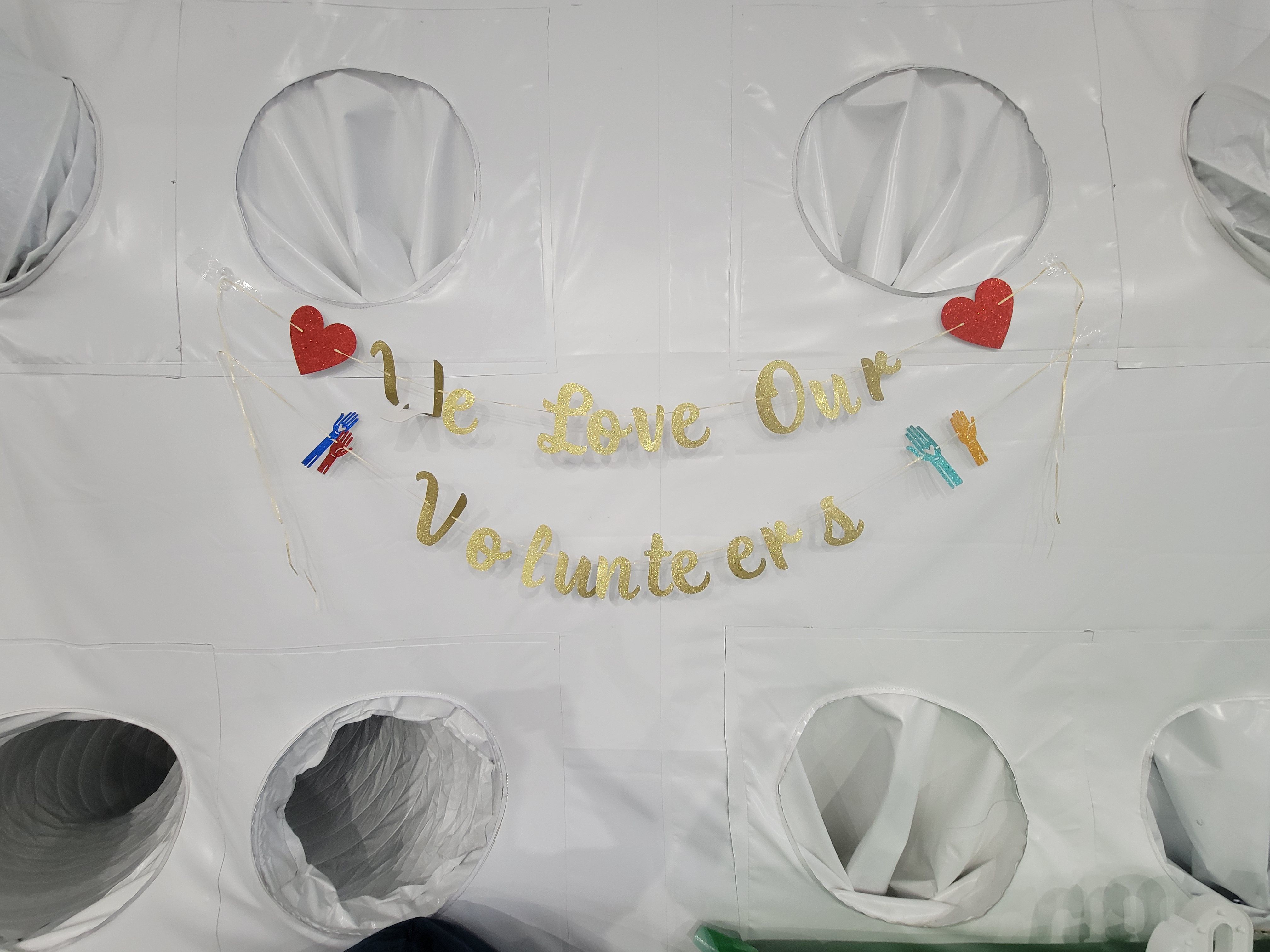 White padded wall with large circular holes; a gold glitter banner with red hearts and multicolored handprint decorations, suggesting a Valentine-themed display.