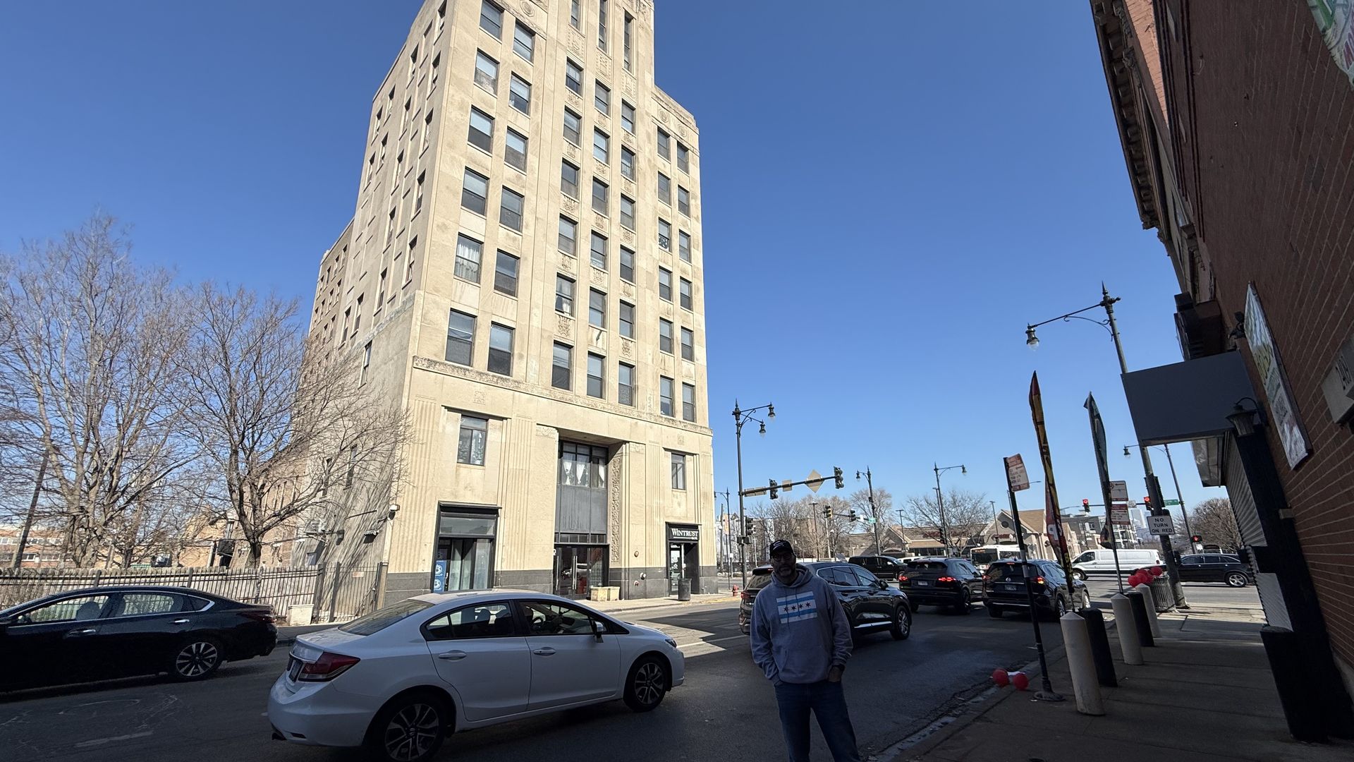 Street view with a tall beige building under clear blue sky, bare trees, cars parked and moving, and a person in a gray hoodie and cap standing on the sidewalk near balloons attached to posts.