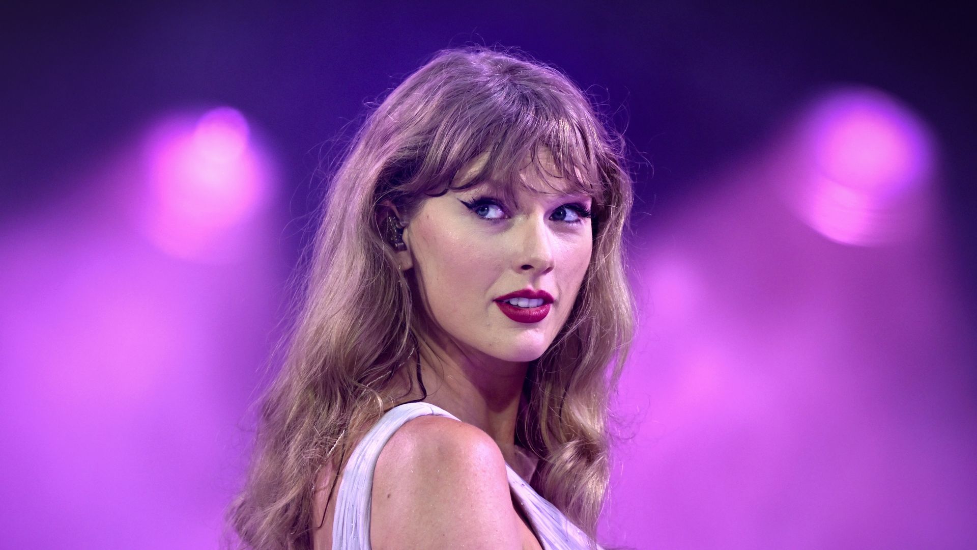 Female performer on a purple-lit stage with long blonde hair and red lipstick, wearing a light sleeveless dress, looking over her shoulder.