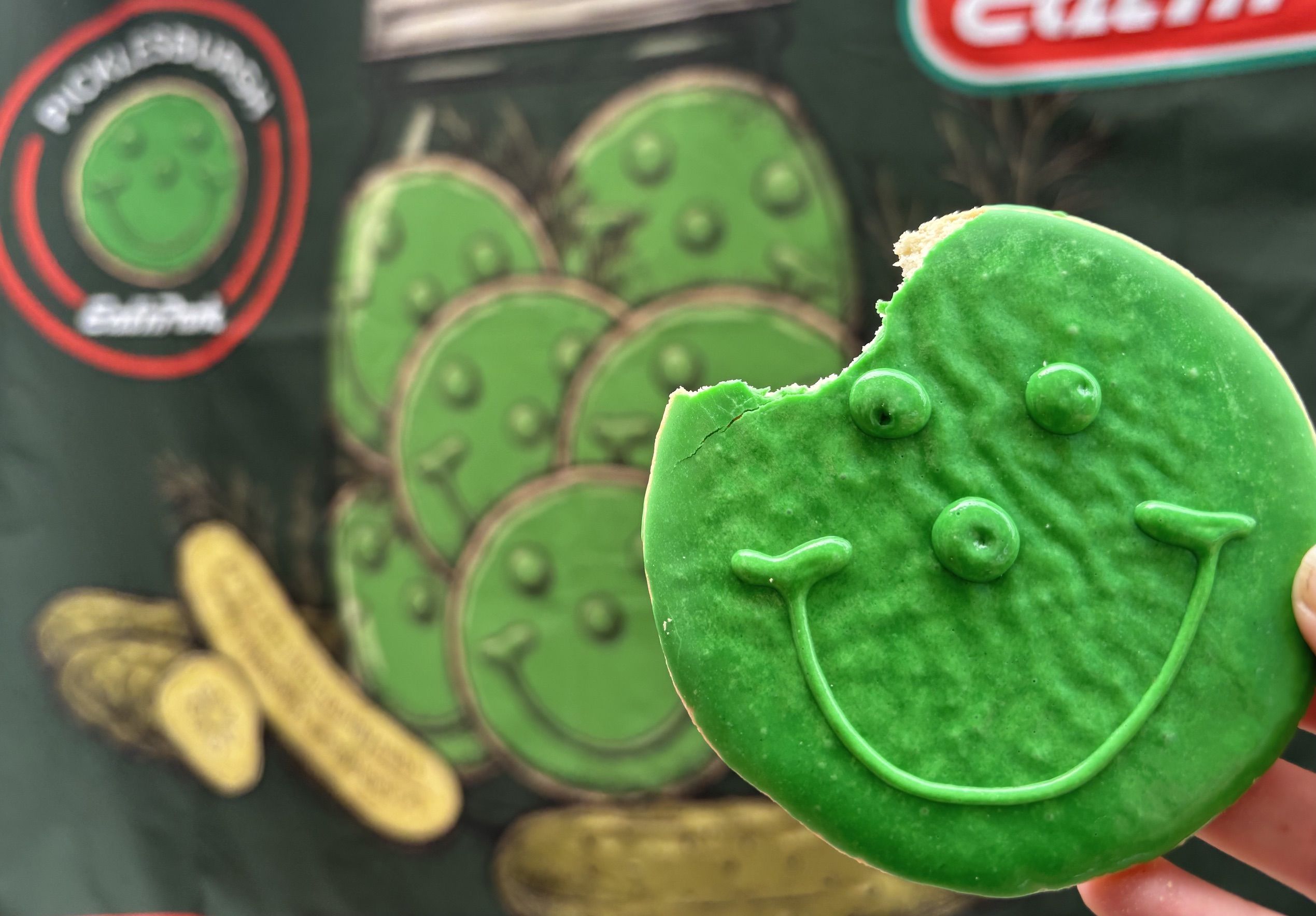 A close-up of a bright green round fondant cookie or cutter being held, with a bite taken from its edge. The background shows blurred green circular shapes and scattered gold coins.