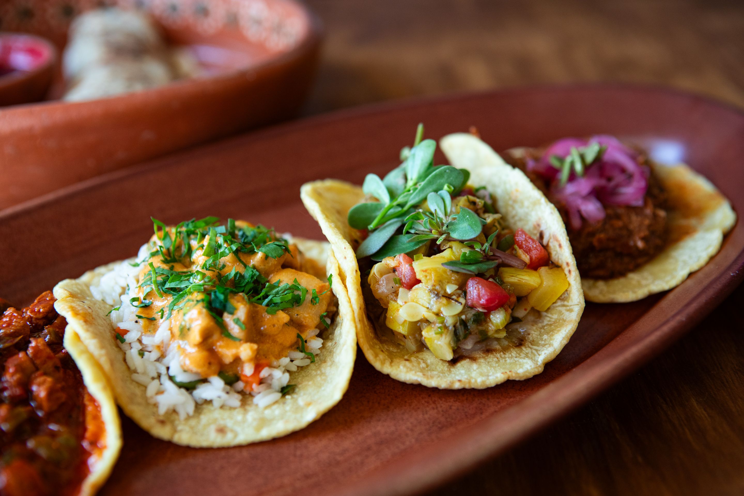 Close-up of three soft tacos on a wooden platter, each filled with colorful toppings: rice and orange sauce with herbs, diced peppers with greens, and shredded meat with red onion.