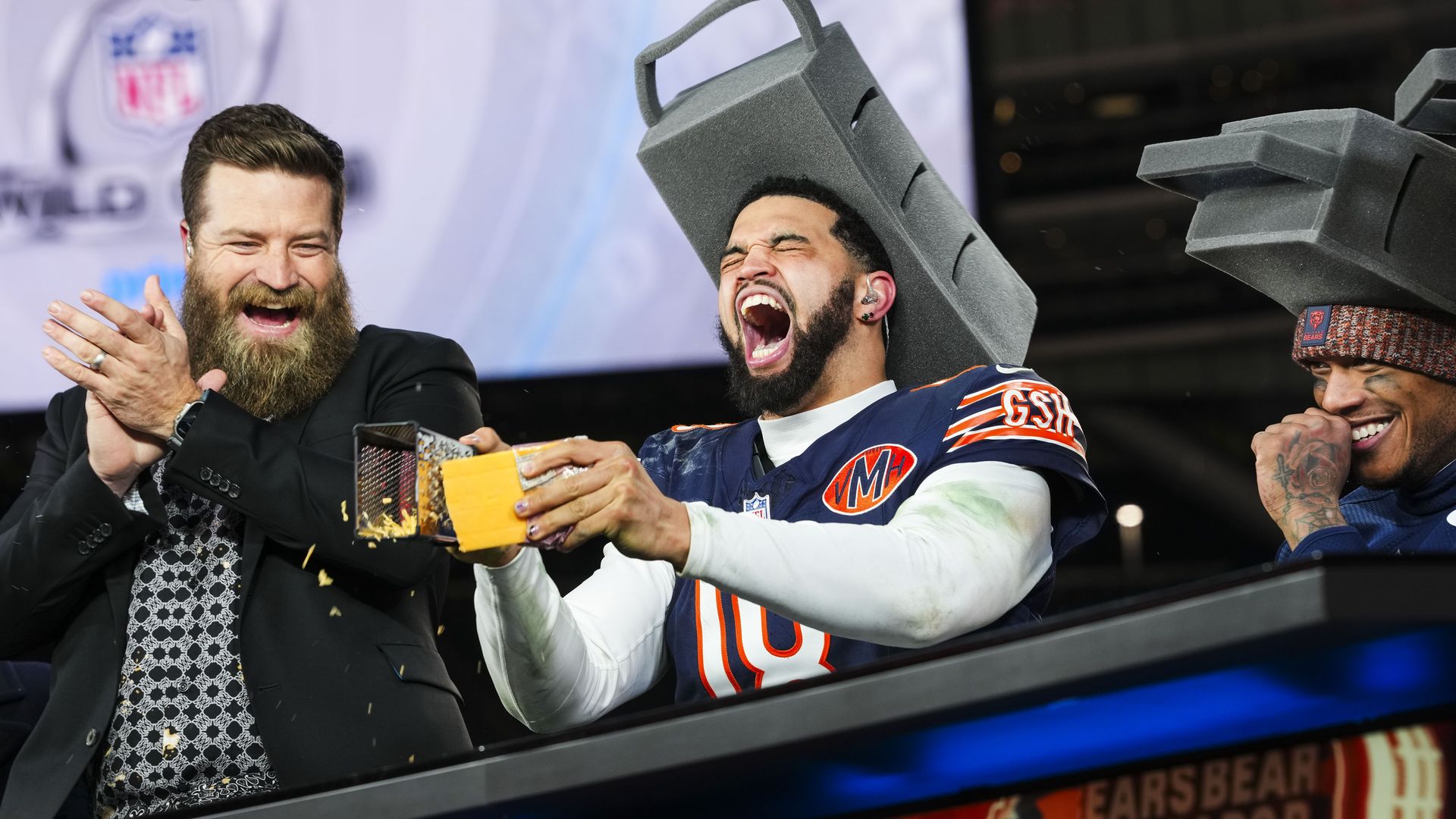 Three excited men on a stadium set; left bearded man in a suit applauds, center a Bears player yells with a foam weight on his head, right a smiling man with a knit cap; BEARS signage in background.