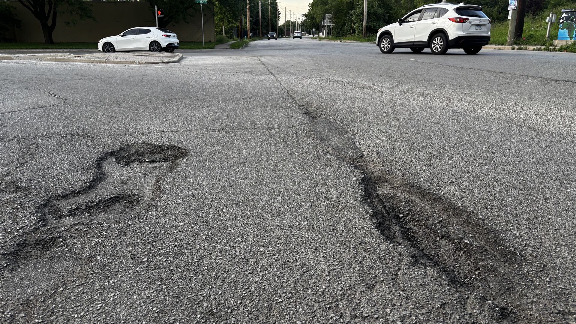 Wide street intersection with rough asphalt, large potholes, and deep cracks. A white sedan is at the left curb and a white SUV sits on the right, with trees and signs in the background.