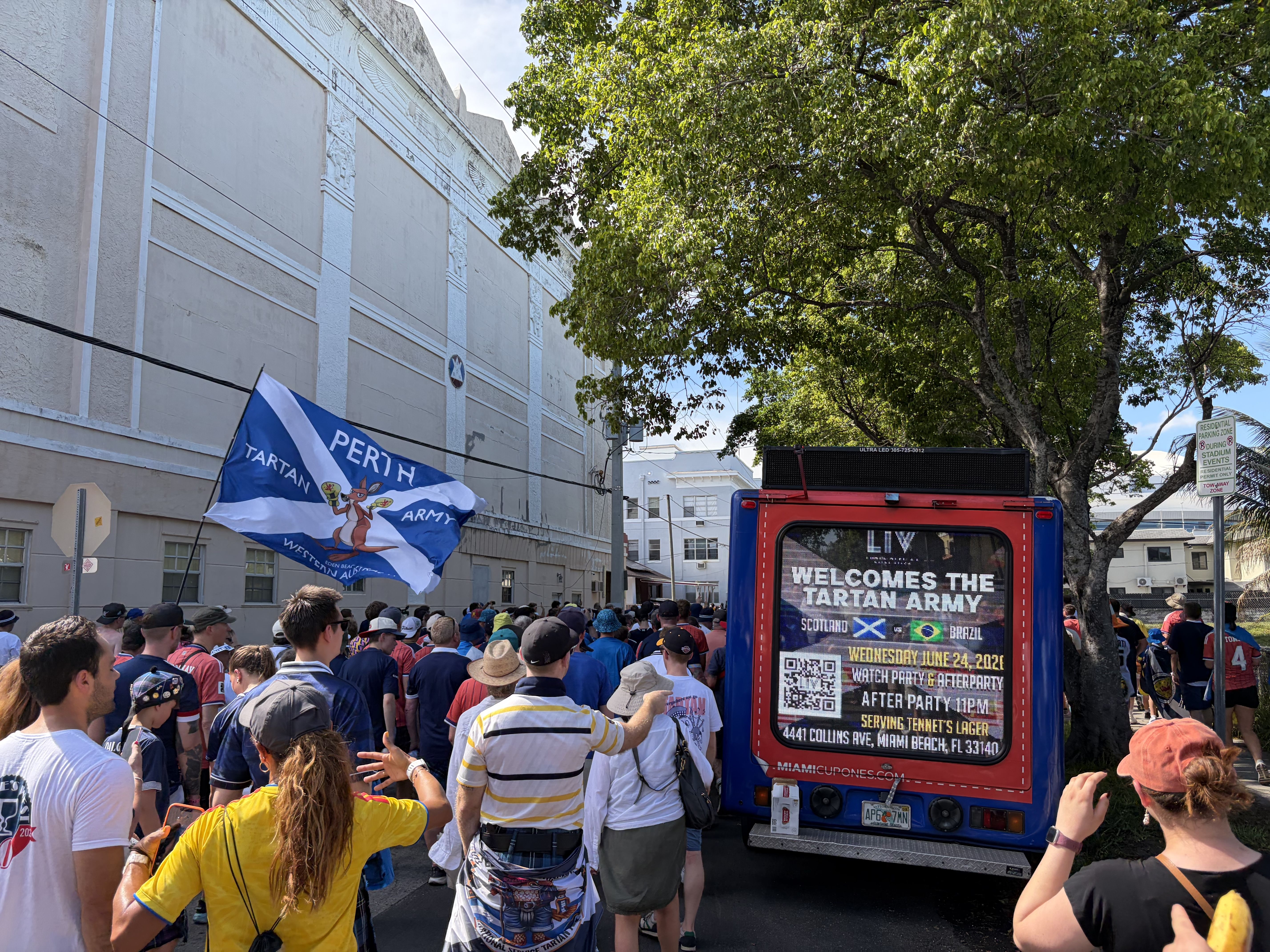 A crowd toting a large Scotland flag marches through a city street past a tree. There's also a video board displaying a message welcoming the Tartan Army.