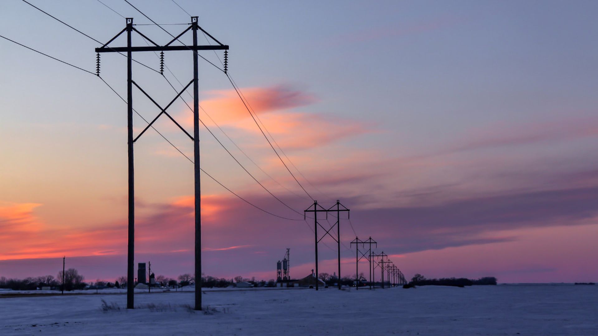 Rows of power lines stretch across a snowy field at sunset, with a sky colored in shades of purple, pink, and orange and silhouettes of distant buildings and trees.