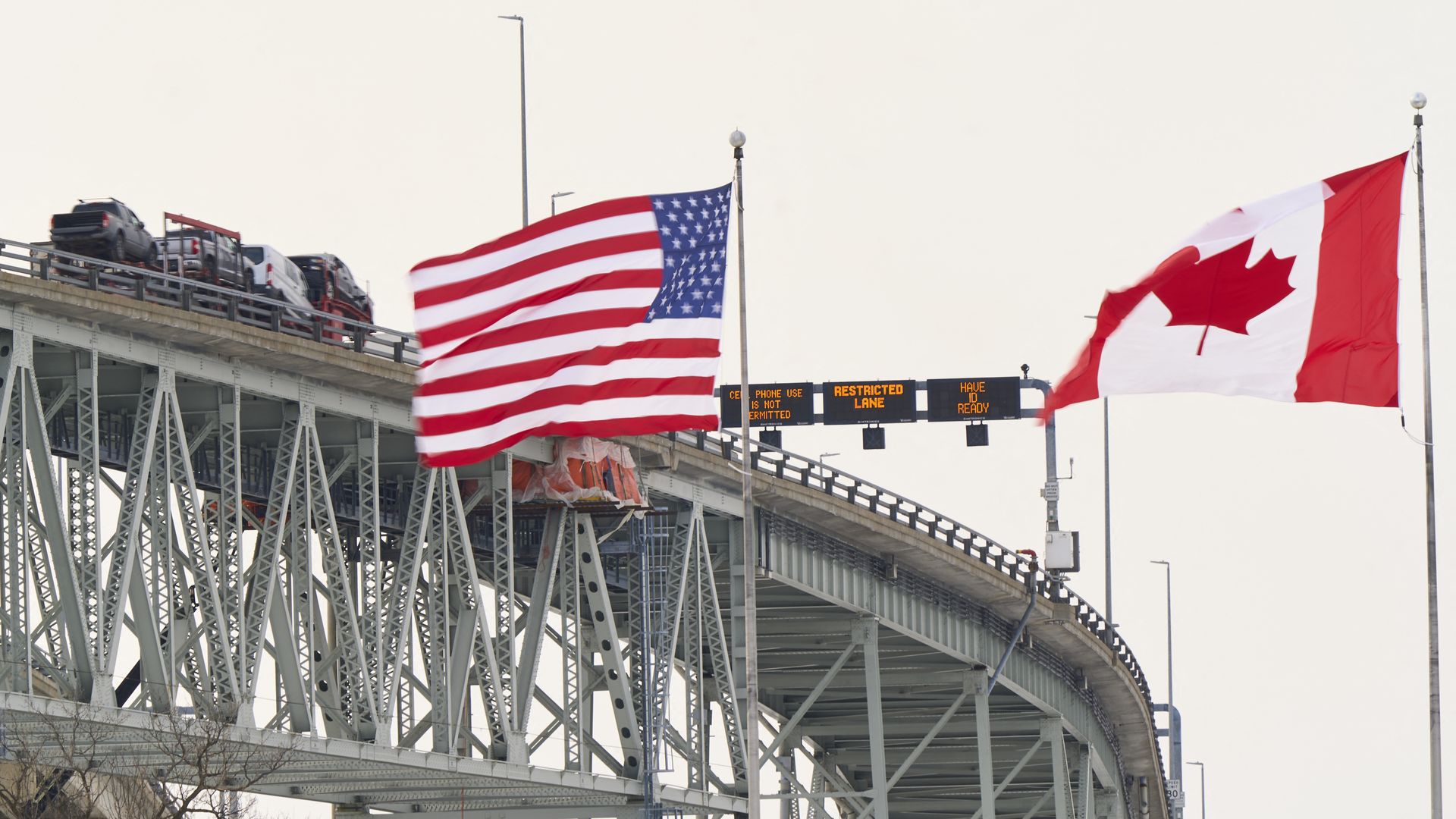 On a metal bridge, American and Canadian flags wave above a roadway with cars; electronic signs read "RESTRICTED LANE" in the background.