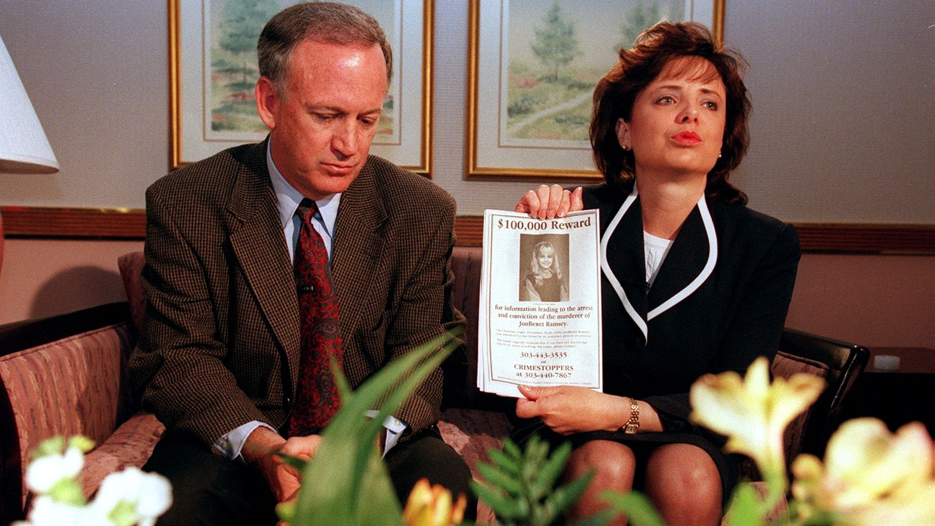 John Ramsey in a brown suit looking down beside Patsy Ramsey in a black suit holding a $100,000 reward flyer for information on JonBenet Ramsey's murderer, with flowers in the foreground.