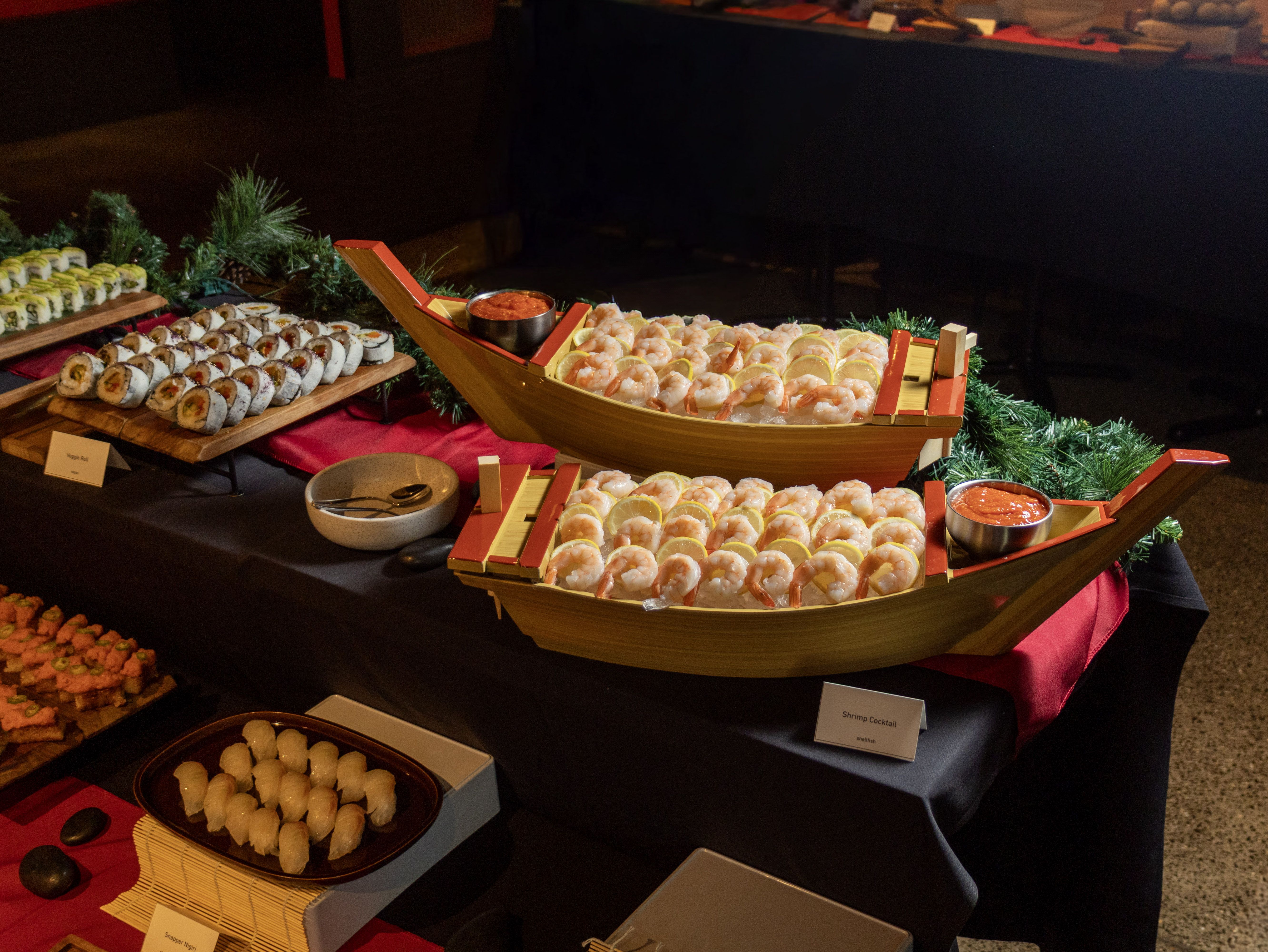 Shrimp cocktail served on ice in two wooden boat-shaped trays, garnished with lemon slices and accompanied by small bowls of red cocktail sauce, displayed on a buffet table.