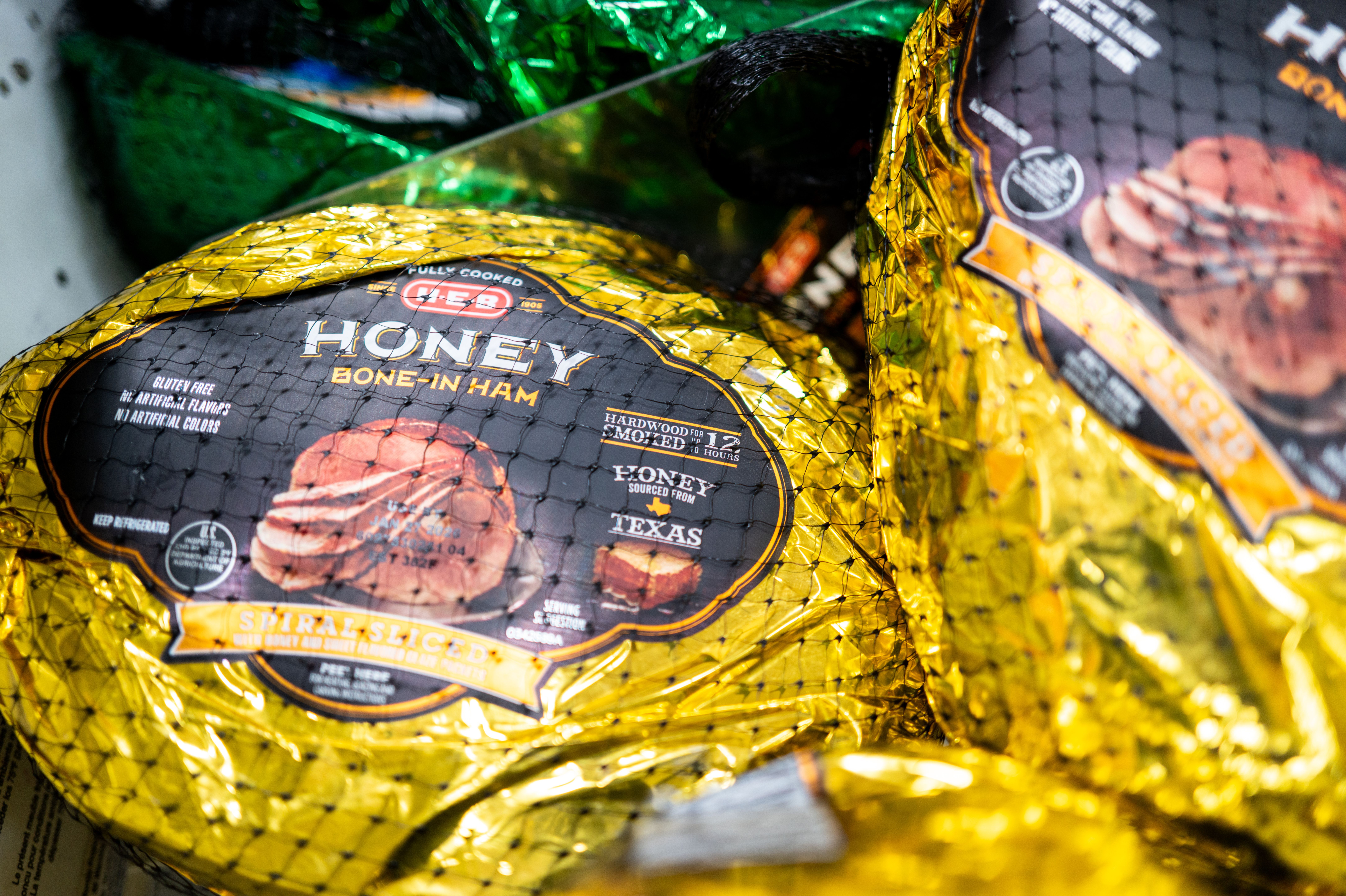 Packaged H-E-B Honey Bone-In Ham wrapped in shiny gold foil with black label, netted for sale, showing spiral sliced ham illustration and Texas honey source.