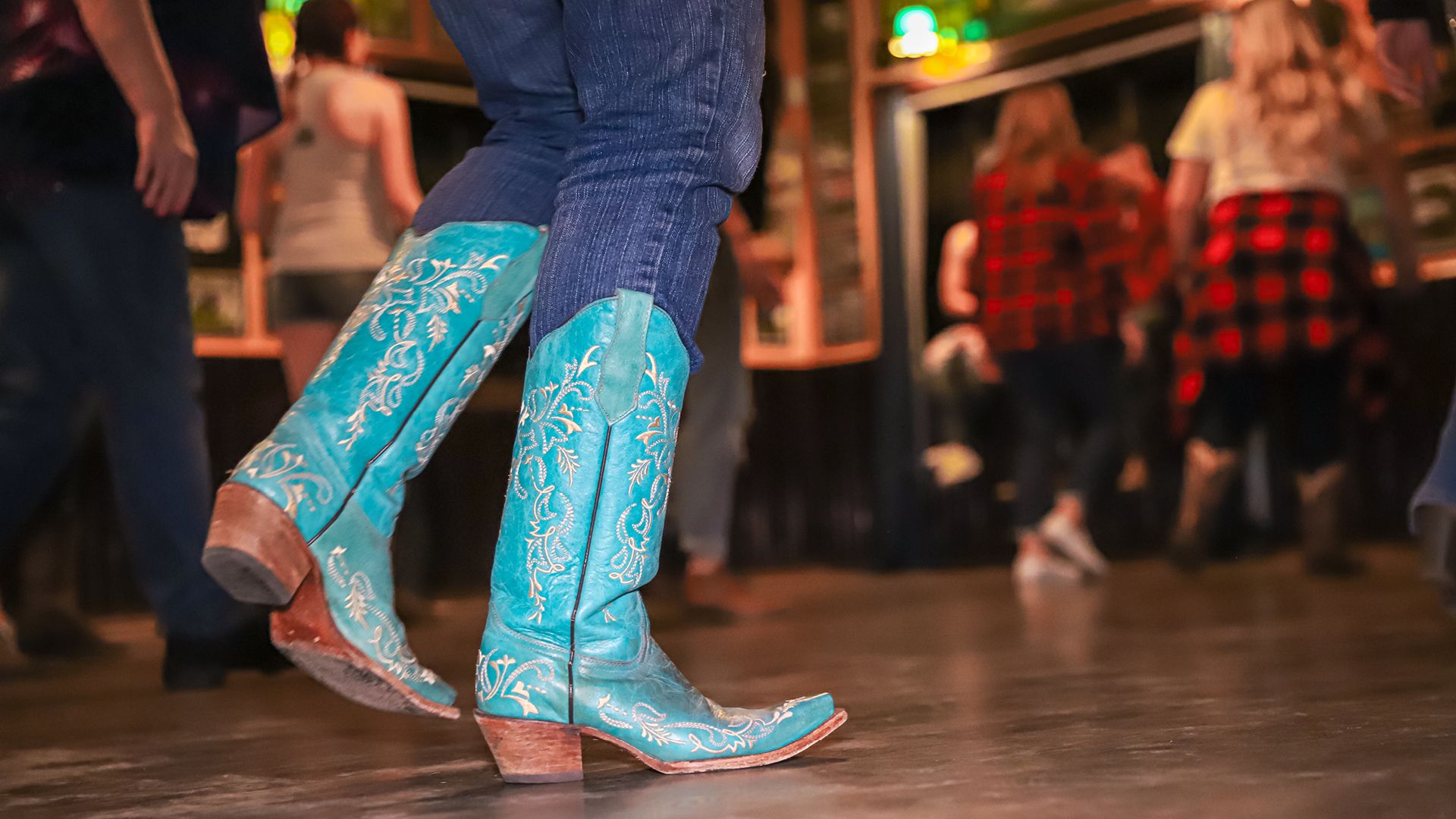 Close-up of a person wearing blue embroidered cowboy boots and jeans dancing on a wooden floor, with people in casual clothing blurred in the background.