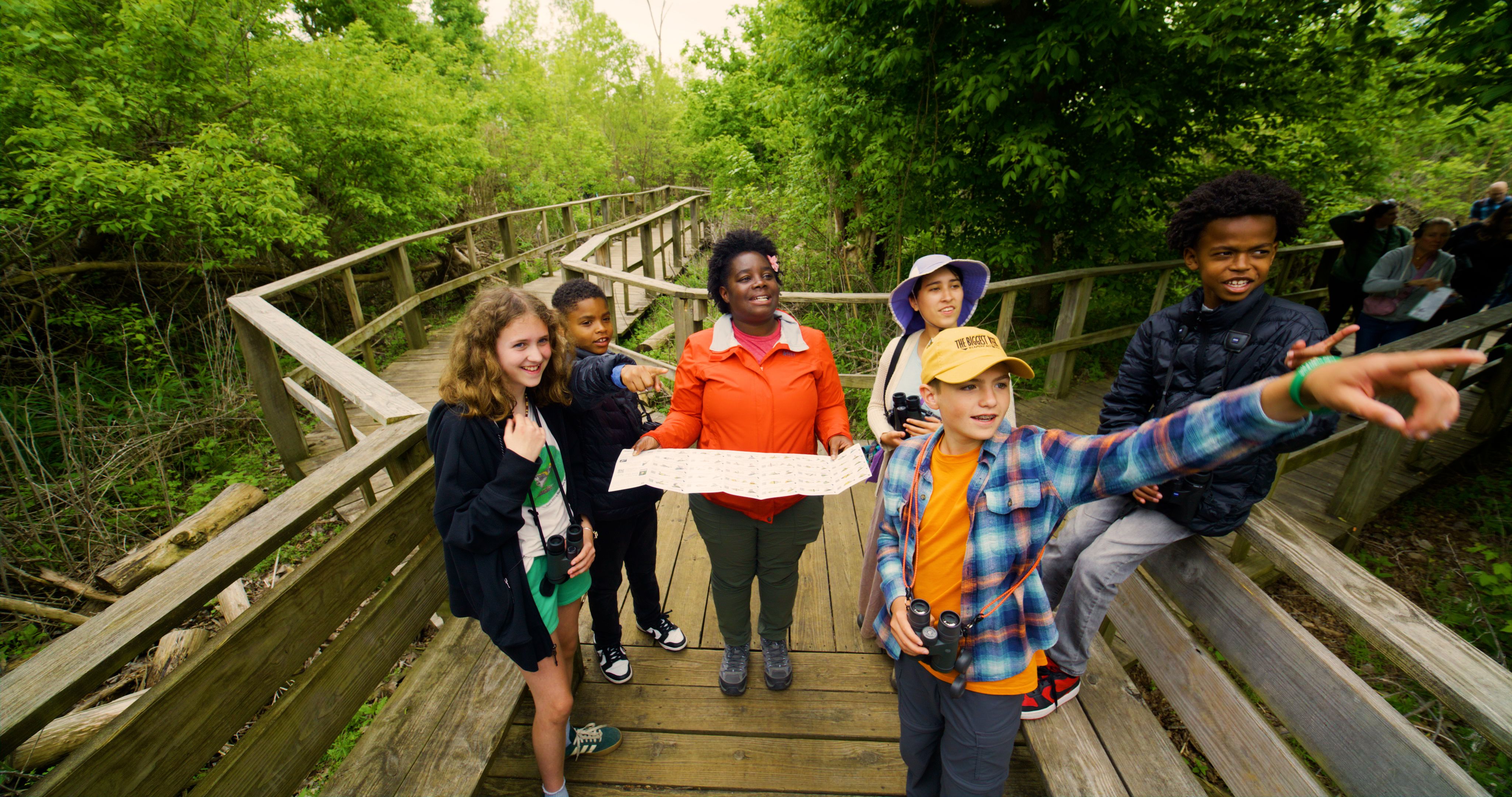 Group of children and a woman with a map on a wooden boardwalk surrounded by green trees. Some kids are pointing and holding binoculars, enjoying an outdoor nature walk.