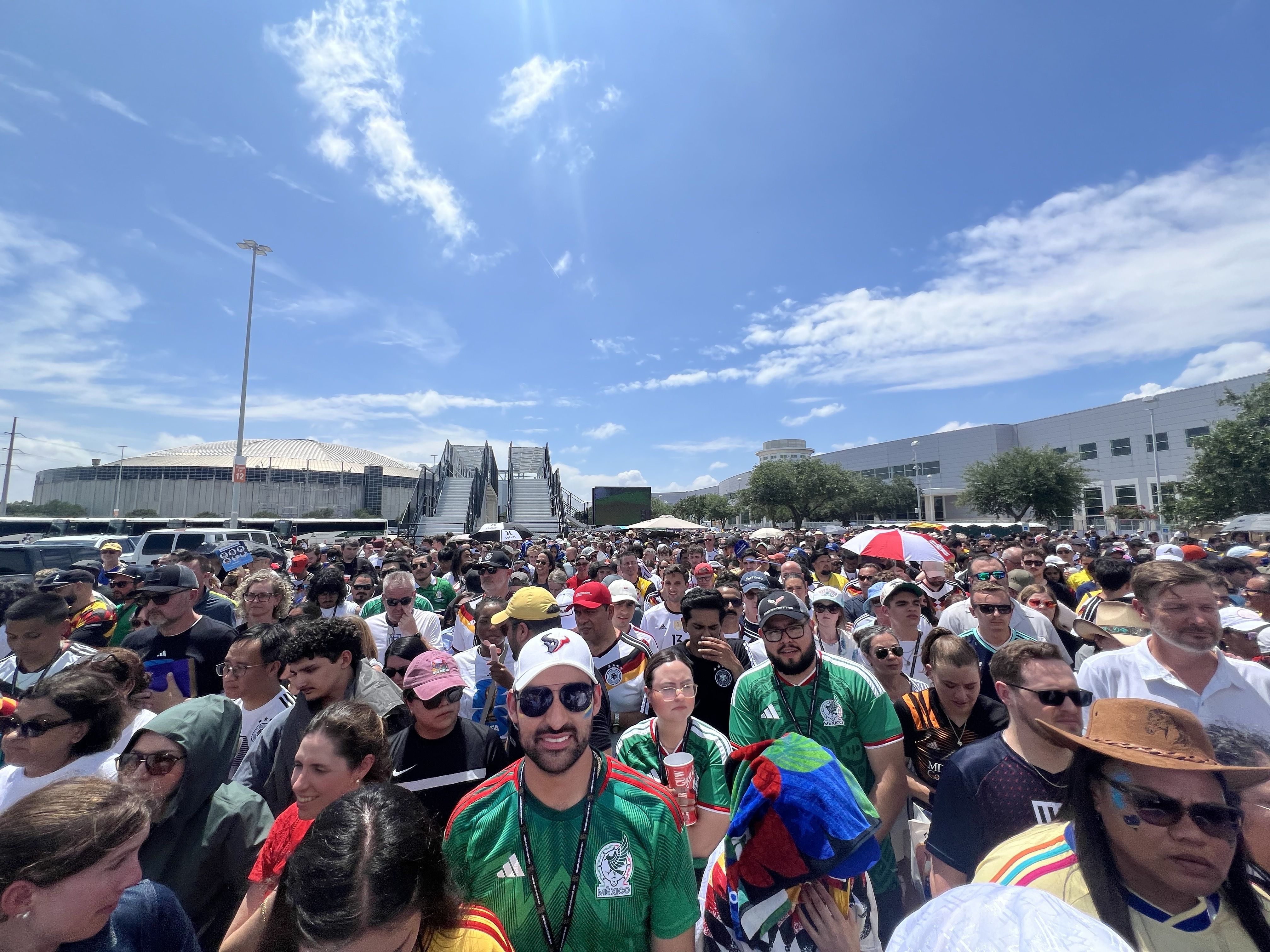 Large crowd of people in colorful jerseys, including green Mexico shirts, gathered outside a stadium under a bright blue sky with scattered white clouds.