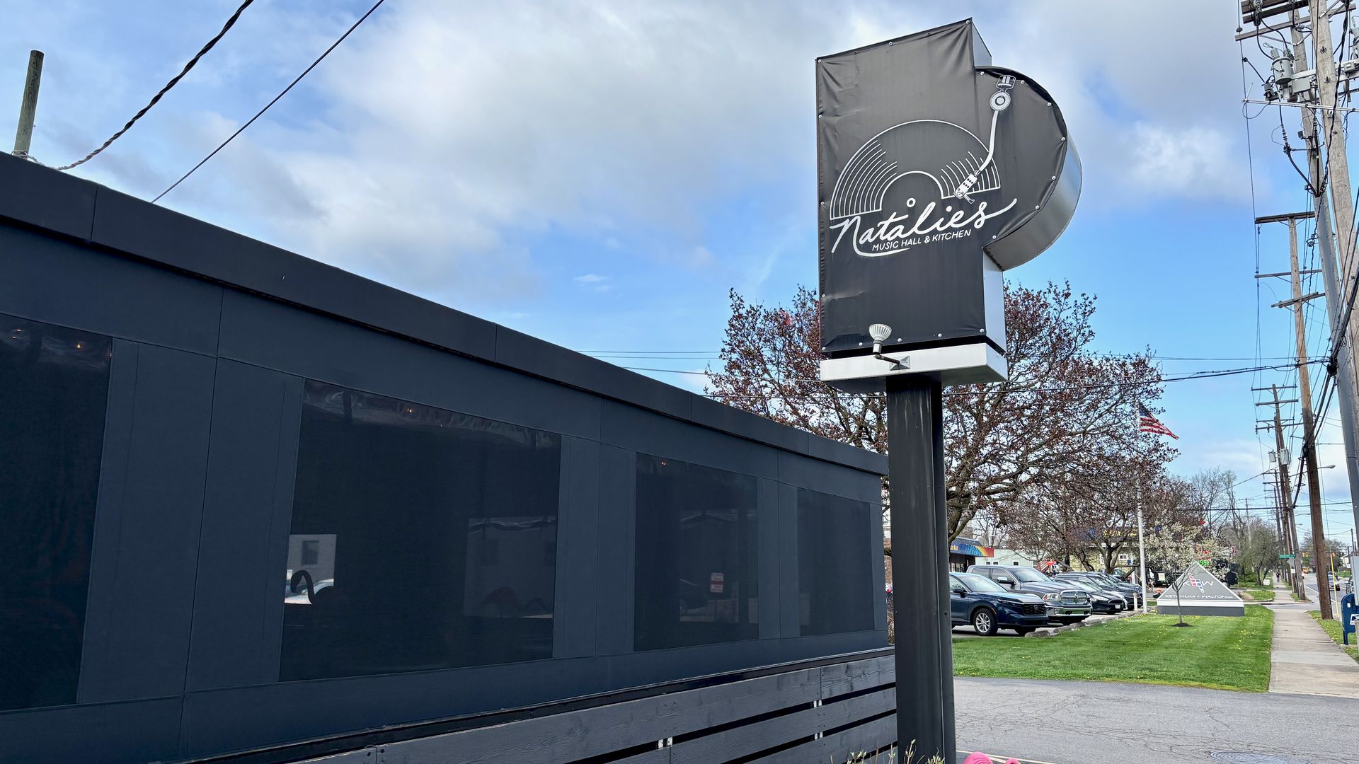 Dark modern storefront with large windows on the left; a tall sign reads "Natalies Music Hall & Kitchen" with a vinyl record graphic. Pink flamingos, parked cars, trees, and a blue sky.