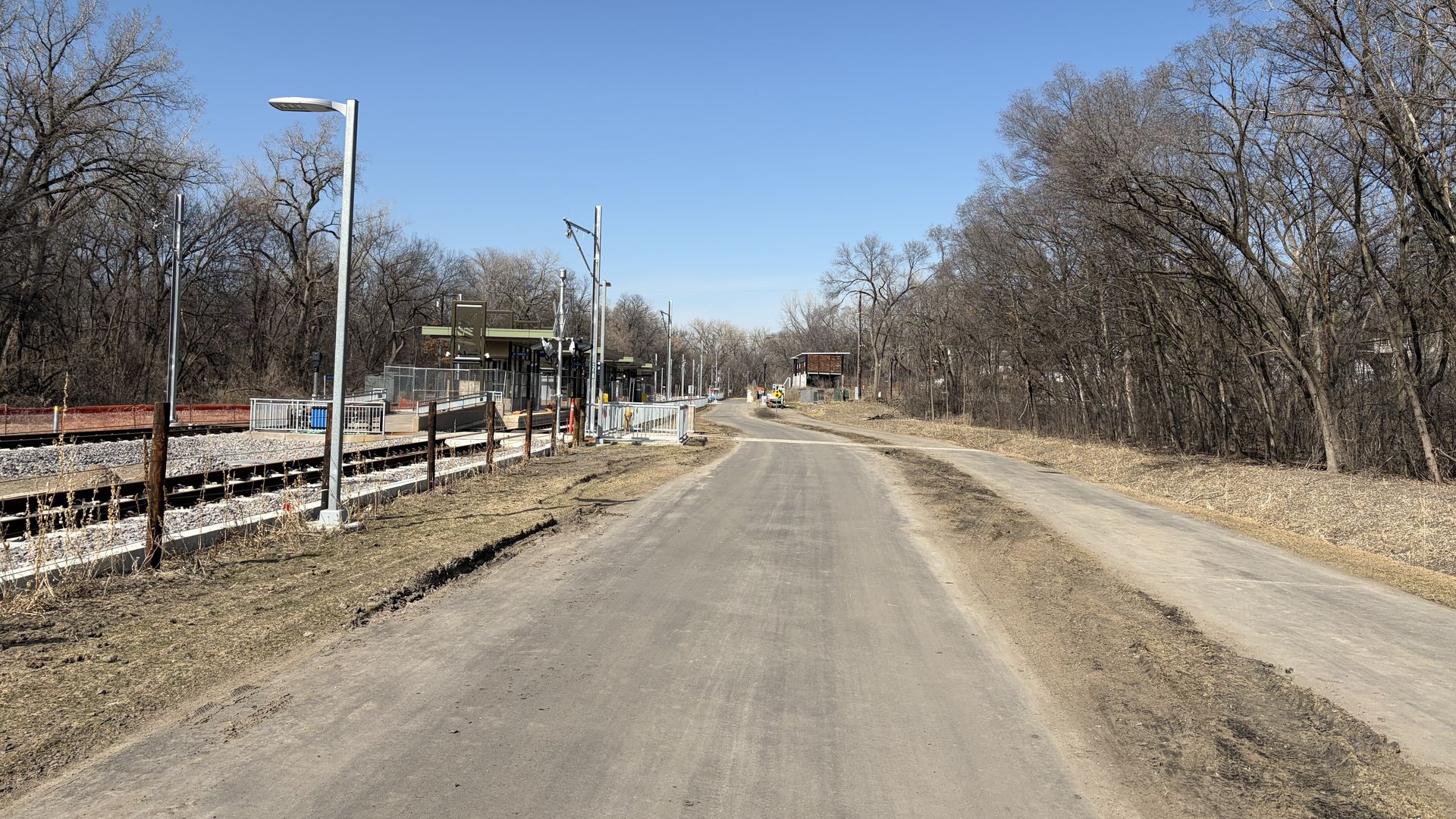 A rural train platform with tracks on the left and a small station building; a dirt road runs center, with a parallel gravel path on the right, flanked by leafless trees under a clear blue sky.