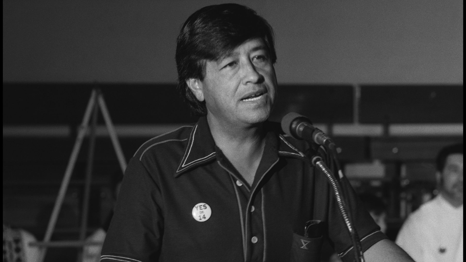A black and white photo of a man speaking into a microphone. He wears a dark-colored shirt with a campaign button that reads "Yes on 14" affixed to the right side. 