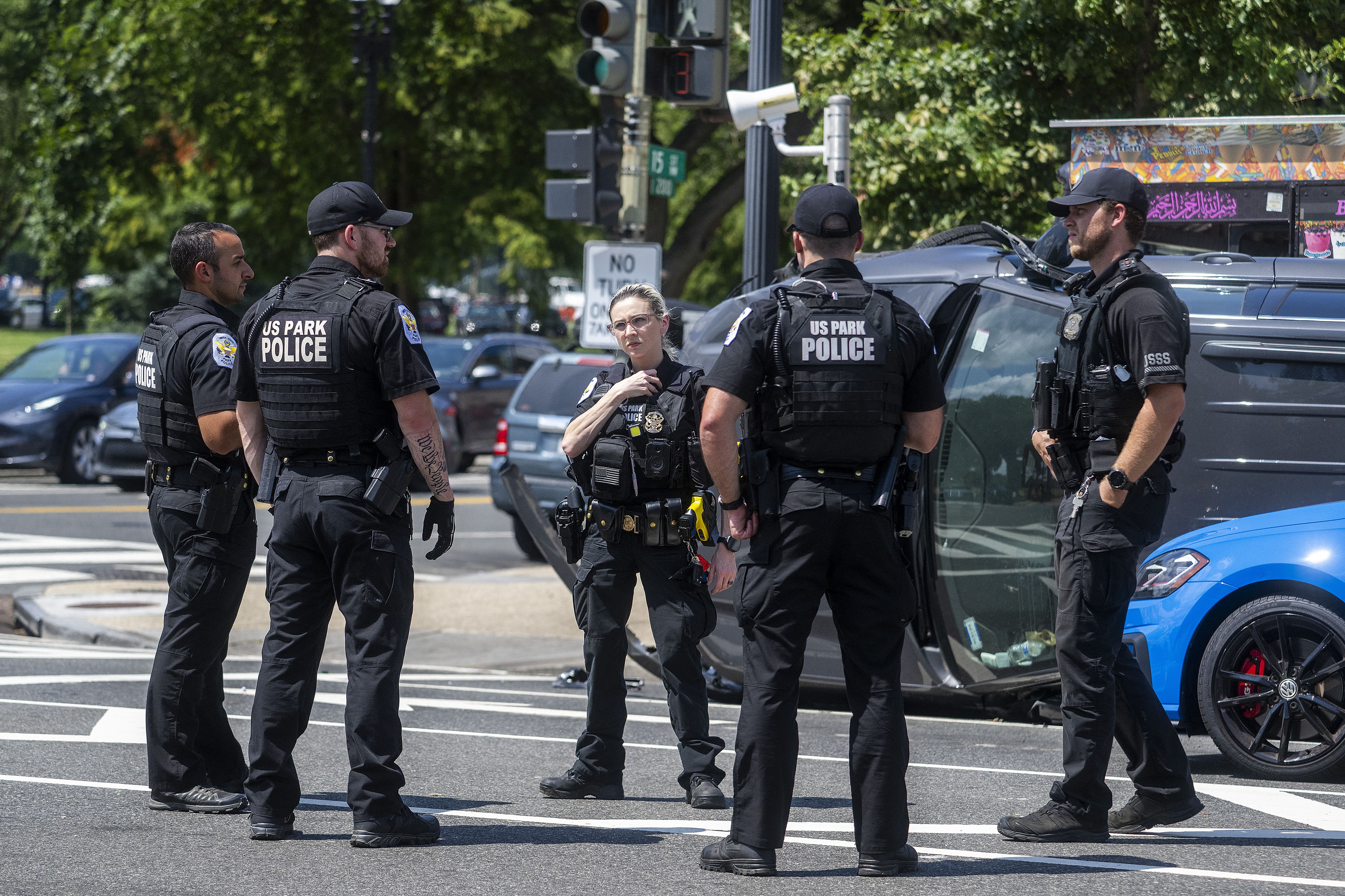 Five US Park Police officers in black uniforms standing on a city street near a flipped dark vehicle and a blue car, with trees and traffic in the background.