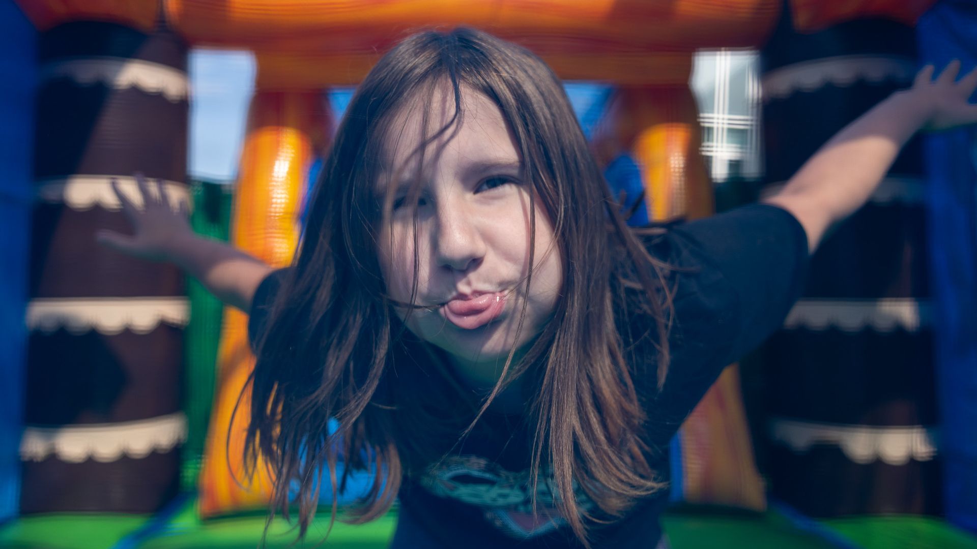 Young girl with long hair smiles through a blurred, colorful inflatable playground. Her arms are raised as orange, blue, and green structures surround her.