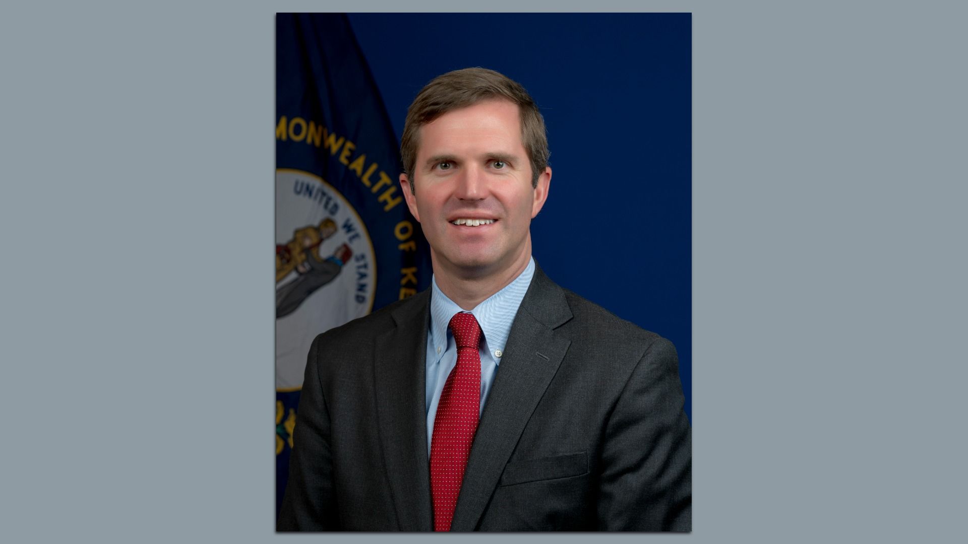 Portrait of a man in a dark suit and red tie, smiling, posed against a dark blue backdrop with part of a seal and flag visible to the left.
