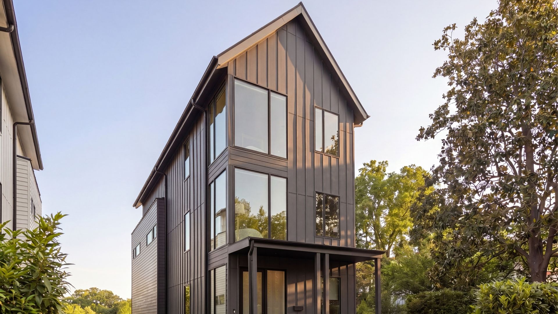 Modern dark-gray house with a tall, sloped roof and expansive glass windows. Front porch seating, lush greenery, and trees on the right under a clear blue sky.