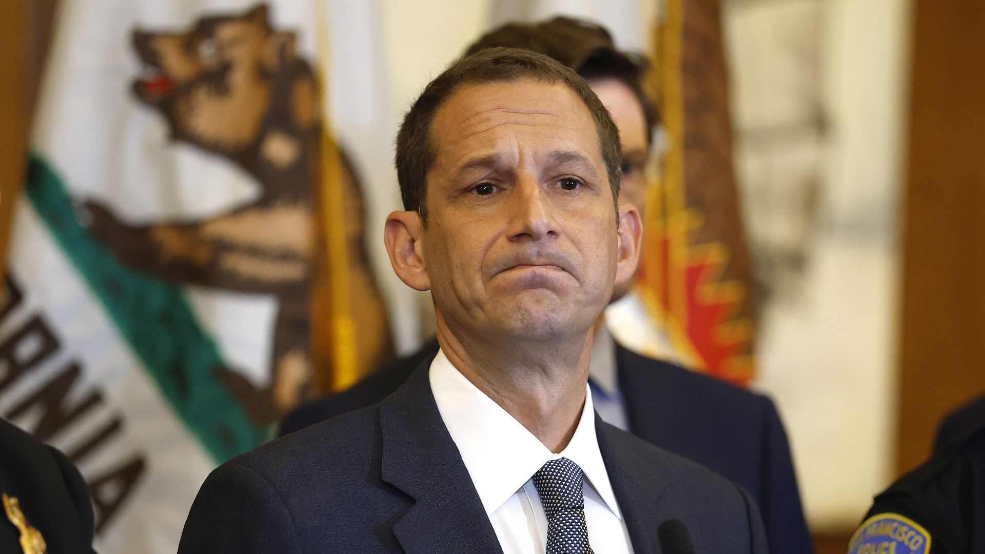 A man in a dark suit and blue patterned tie stands solemnly at a microphone with a California state flag behind him and police officers partially visible.