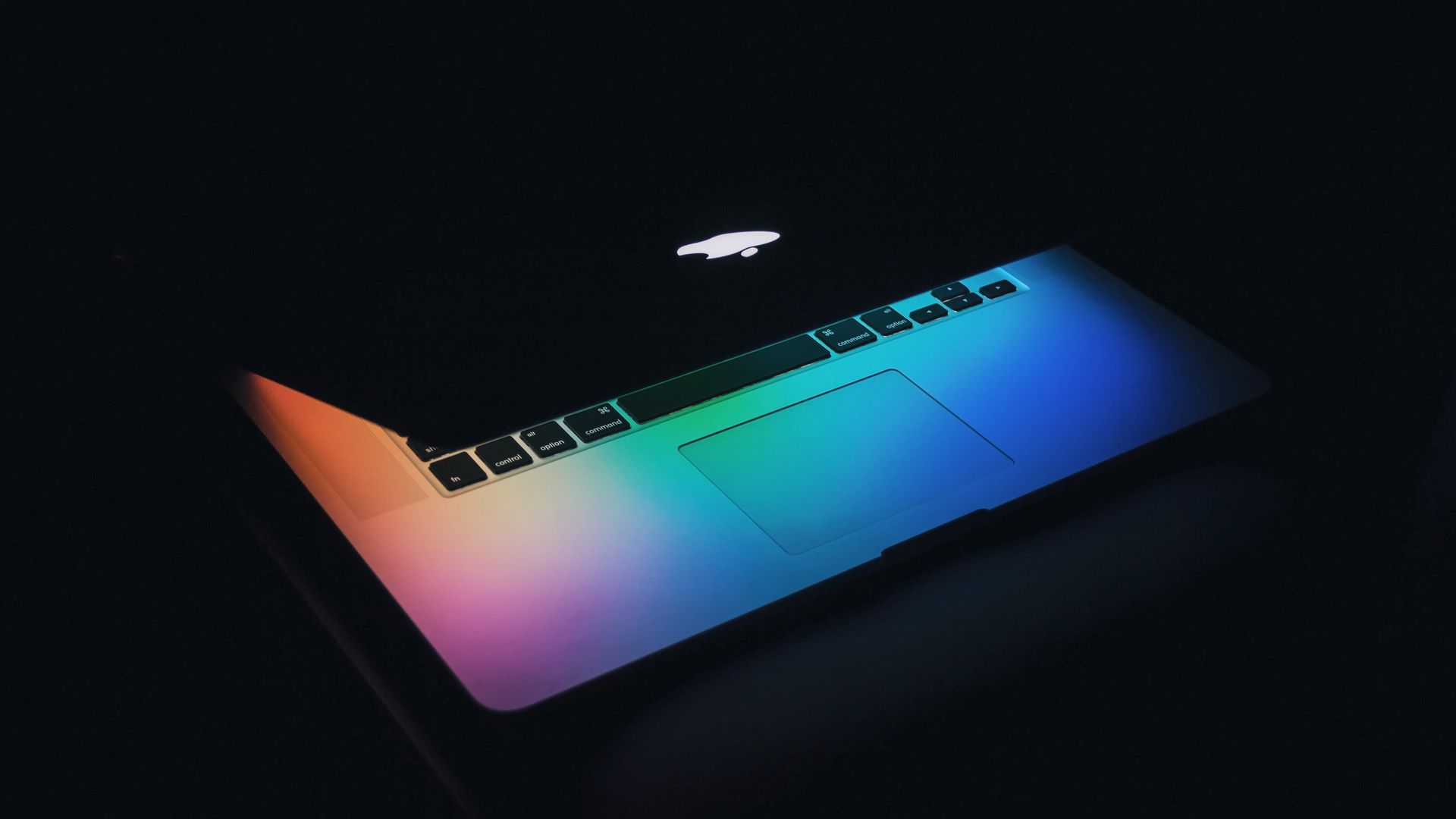 The screen of a partially closed Apple laptop shines with a rainbow glow.