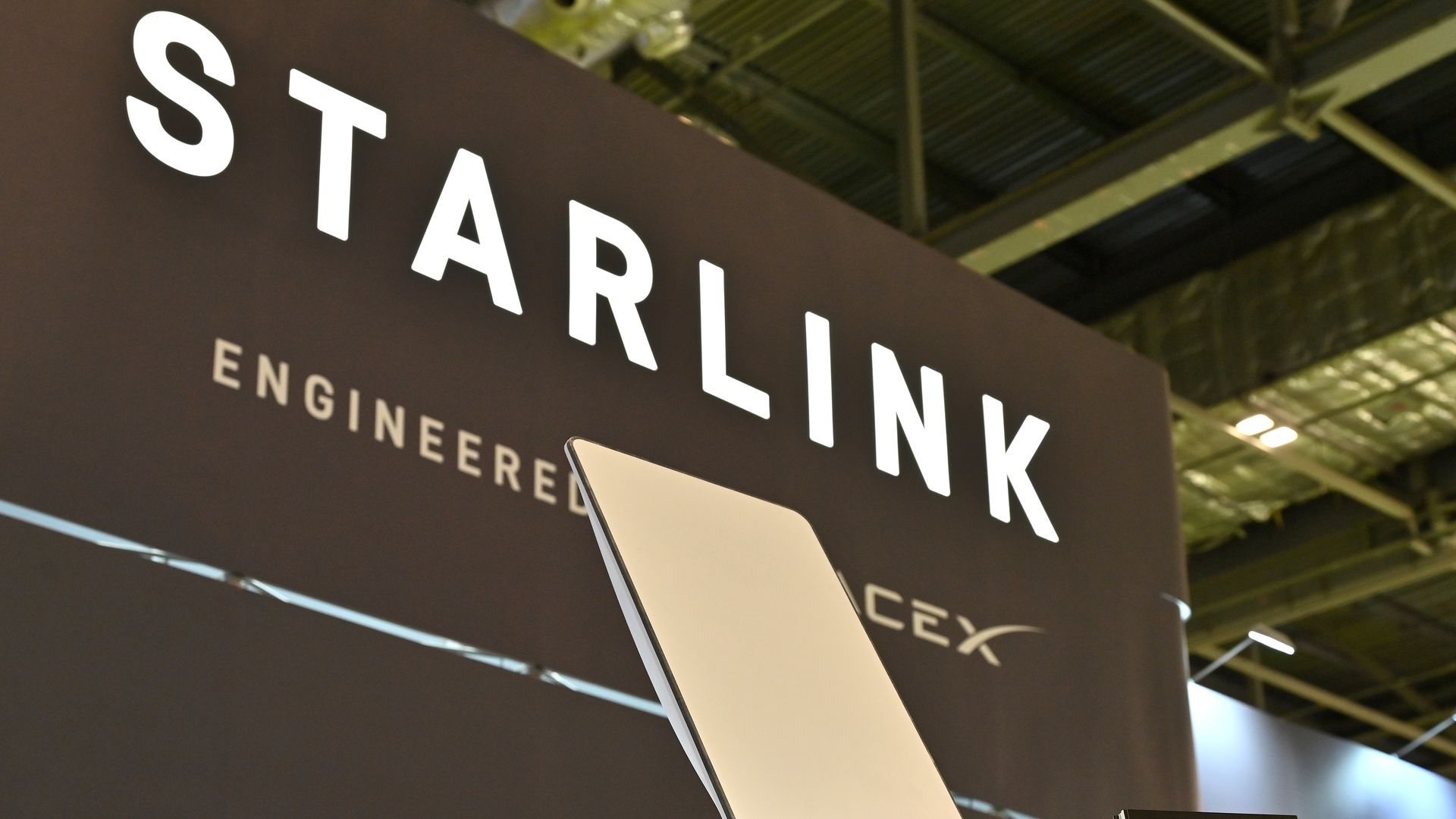 A sleek white Starlink satellite dish displayed on a black stand at an exhibition booth with a large "STARLINK" sign and pricing information. Industrial ceiling visible above.