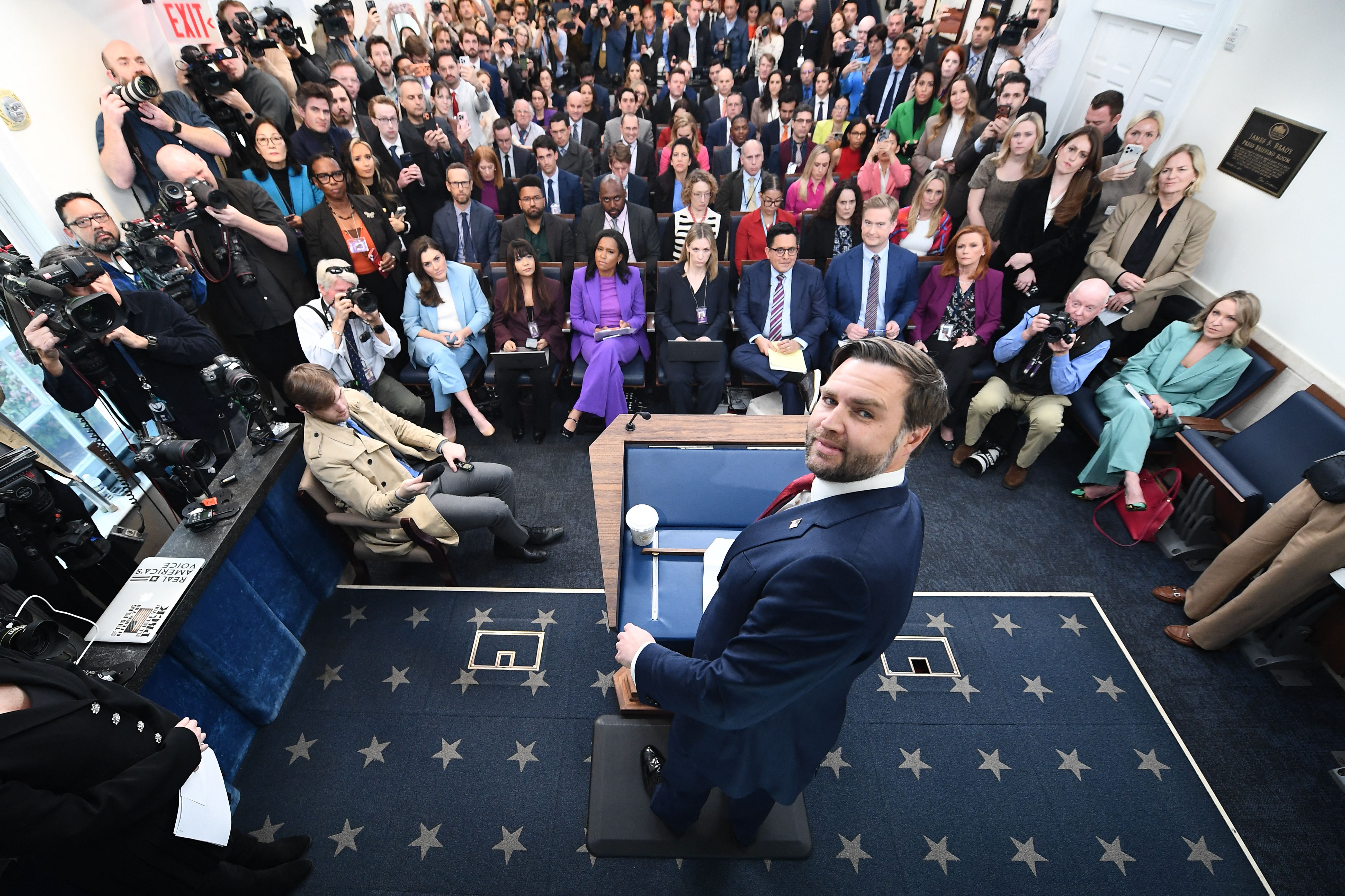 A high-angle shot of Vance briefing a room full of reporters