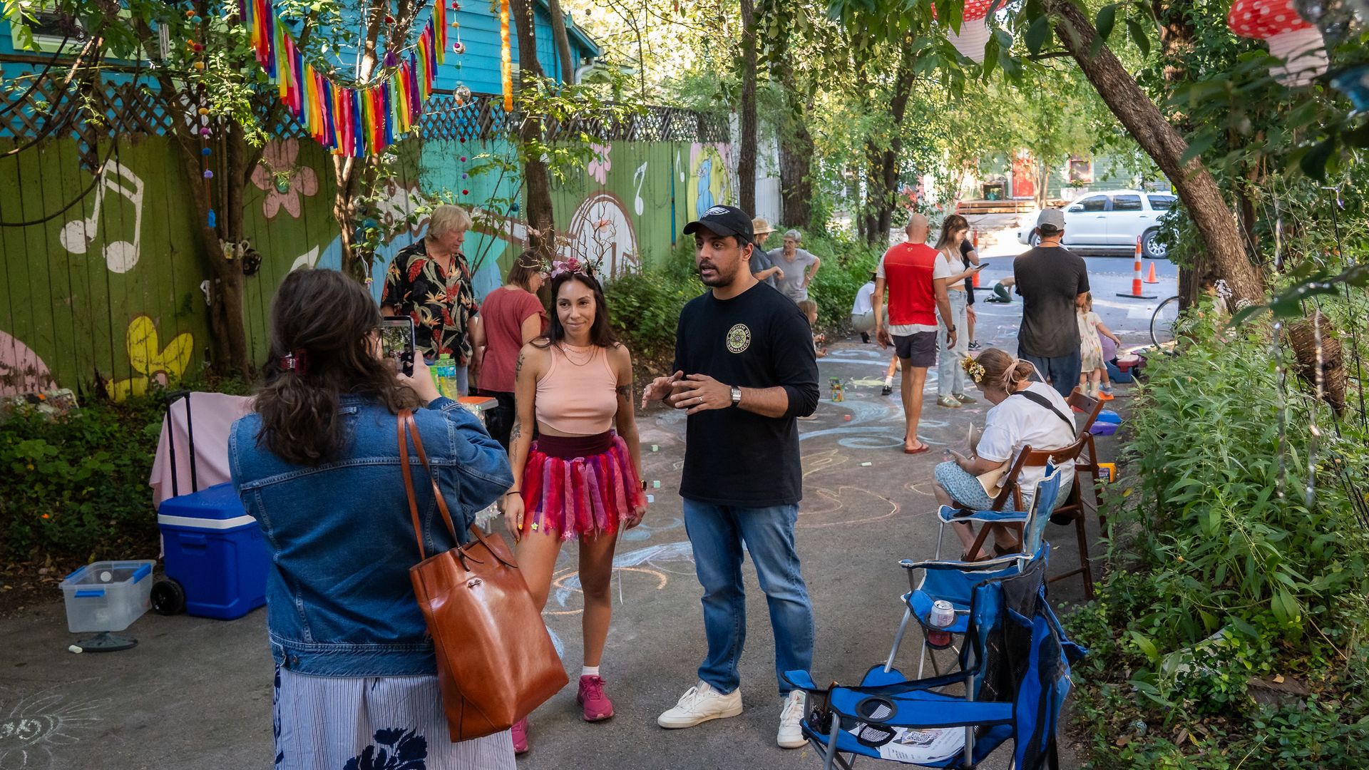 Sunny alley party with colorful bunting and mural walls. A woman in peach and a pink tutu chats with a man in black as a bystander films; chalk art covers the pavement, trees and a blue house nearby.