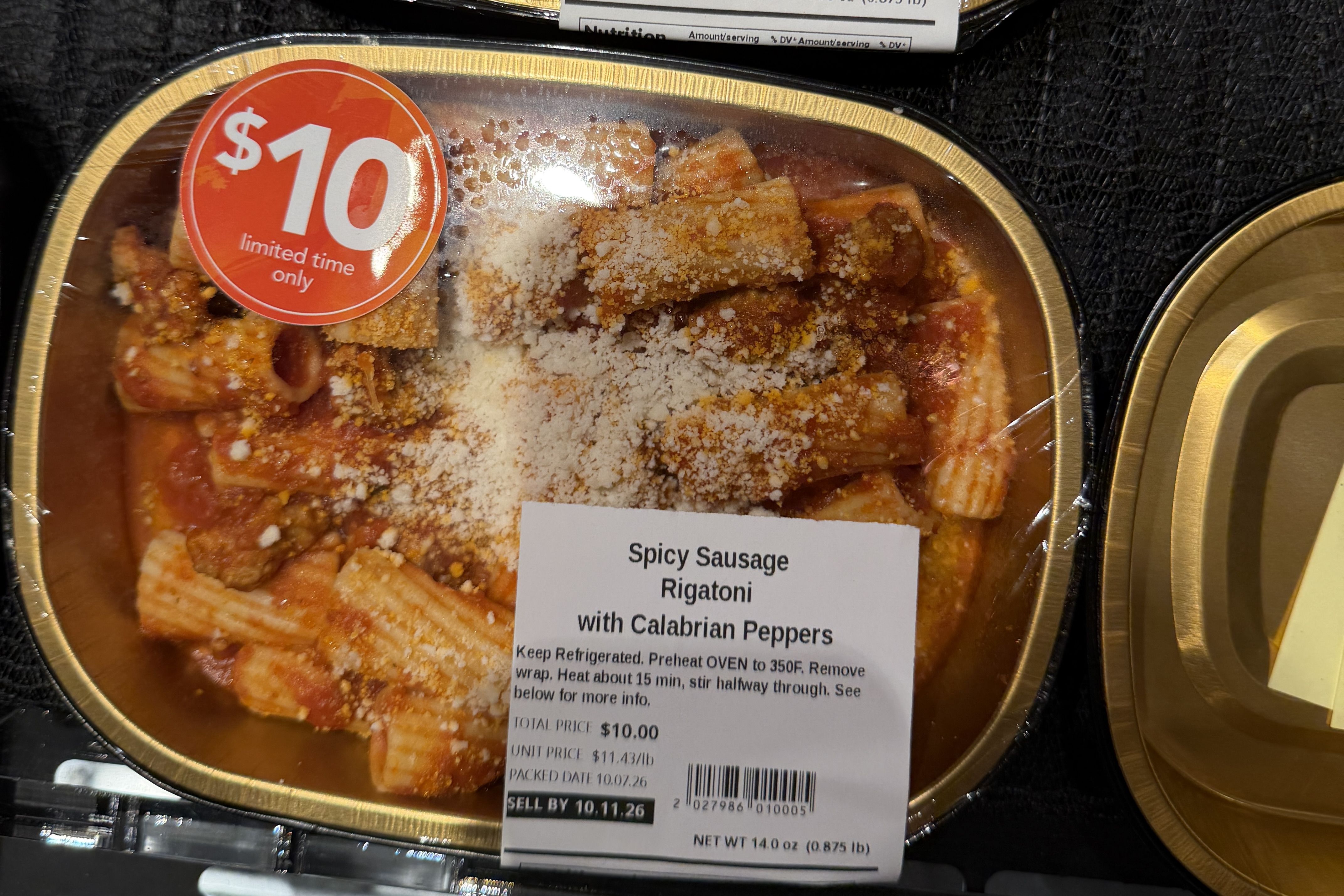 Takeout container of spicy sausage rigatoni in tomato sauce, topped with grated cheese. A red $10 "limited time only" sticker sits on the lid; label reads "Spicy Sausage Rigatoni with Calabrian Peppers".