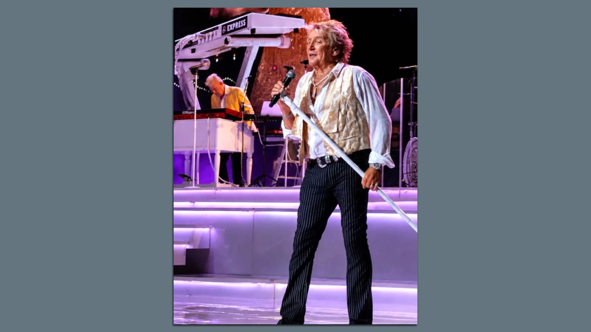 Male singer with blond hair, wearing a cream vest and white shirt, striped pants, holding a microphone and white stand on a lit stage; keyboardist in yellow in the background.