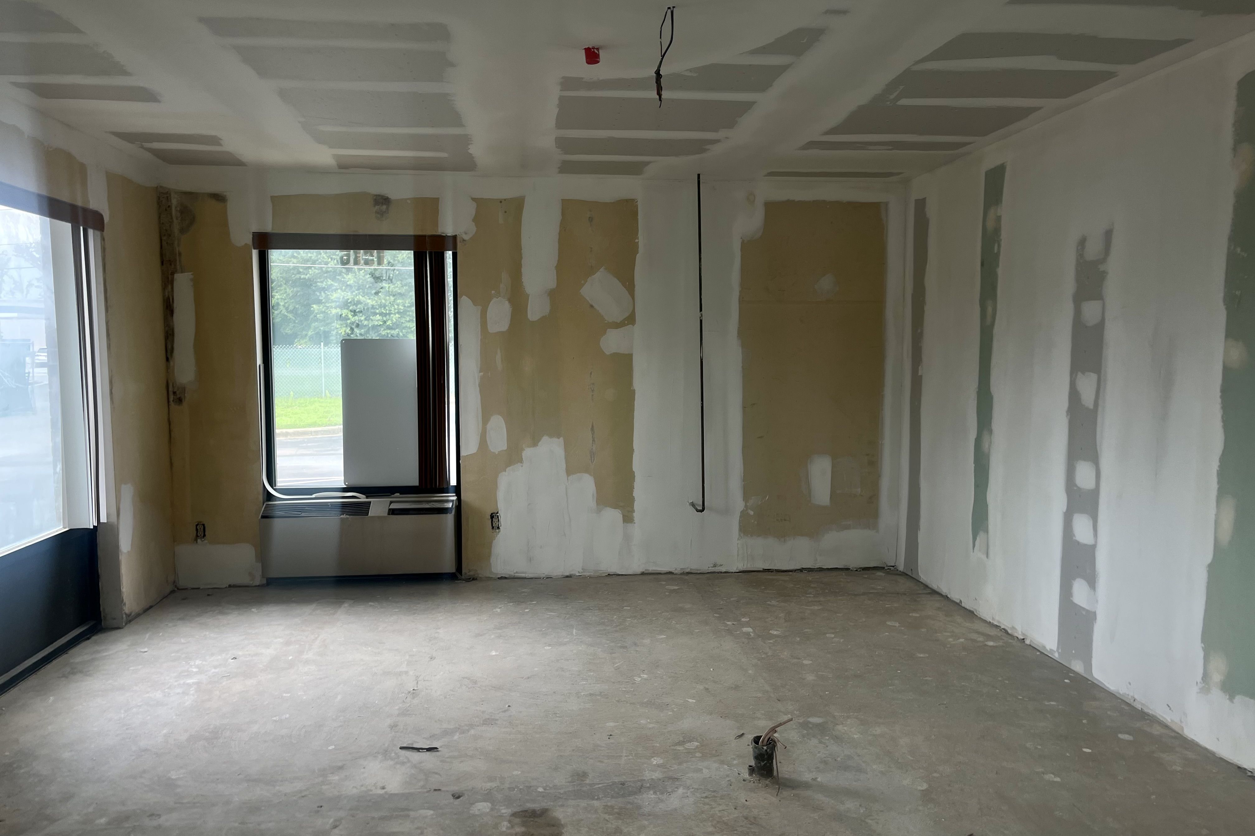 Empty room with unfinished drywall and patches, bare concrete floor, one window with a metal heater below, and exposed wiring hanging from the ceiling. Light from outside visible.