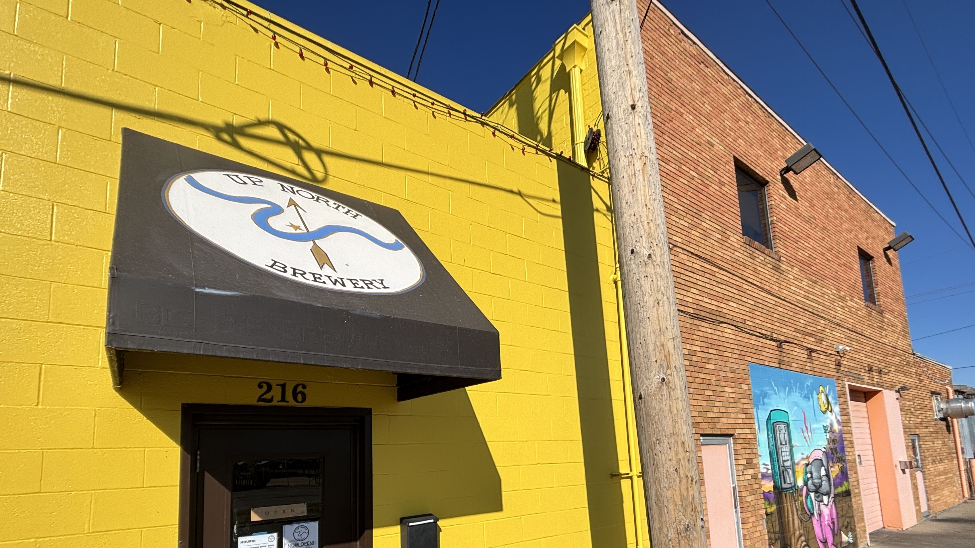 Bright yellow building with a black awning reading "Up North Brewery" above door number 216; adjacent red brick building features colorful mural and utility pole in front under clear blue sky.