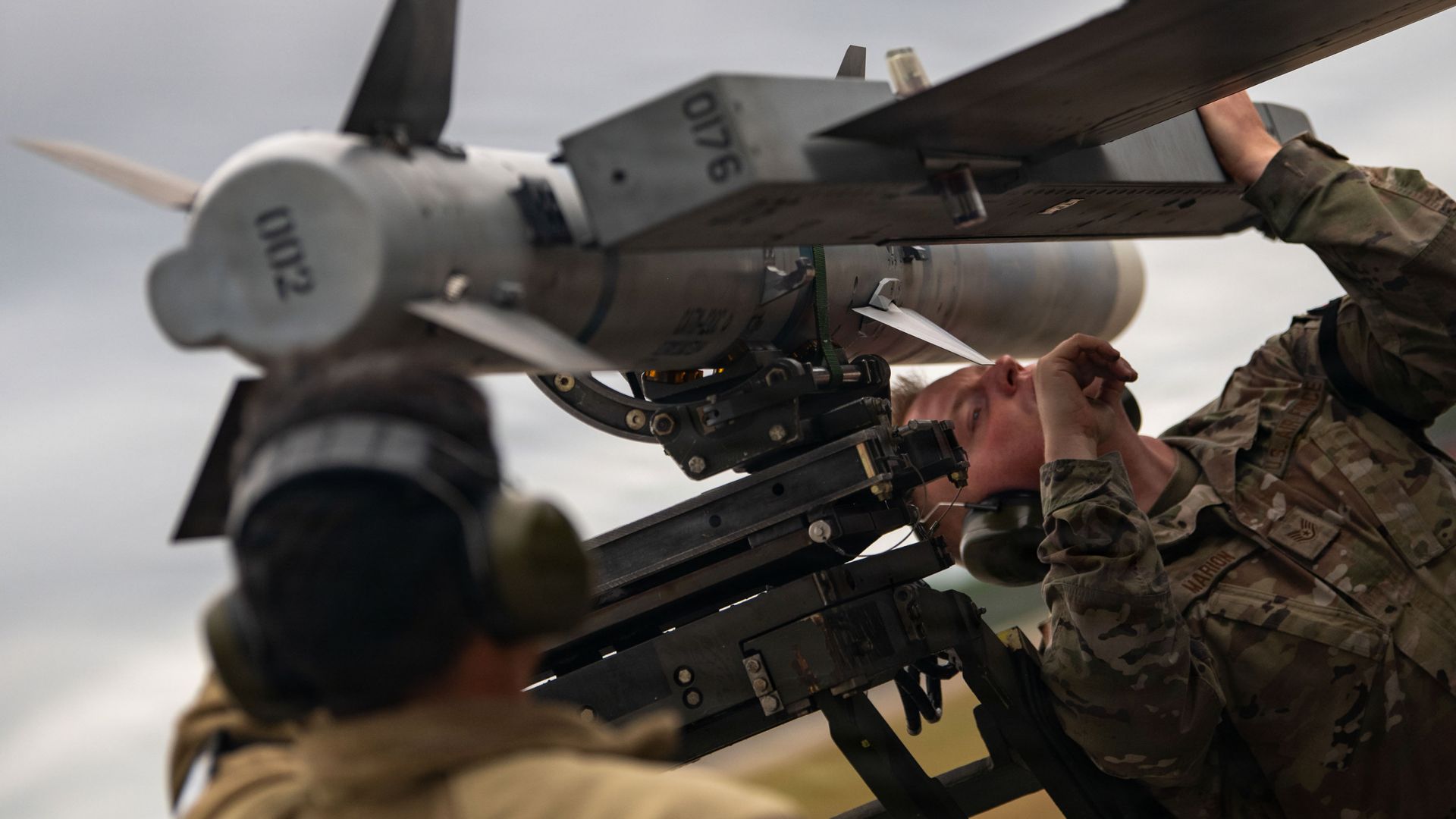 Two military personnel in camouflage uniforms working with a large mounted missile against a cloudy sky background.
