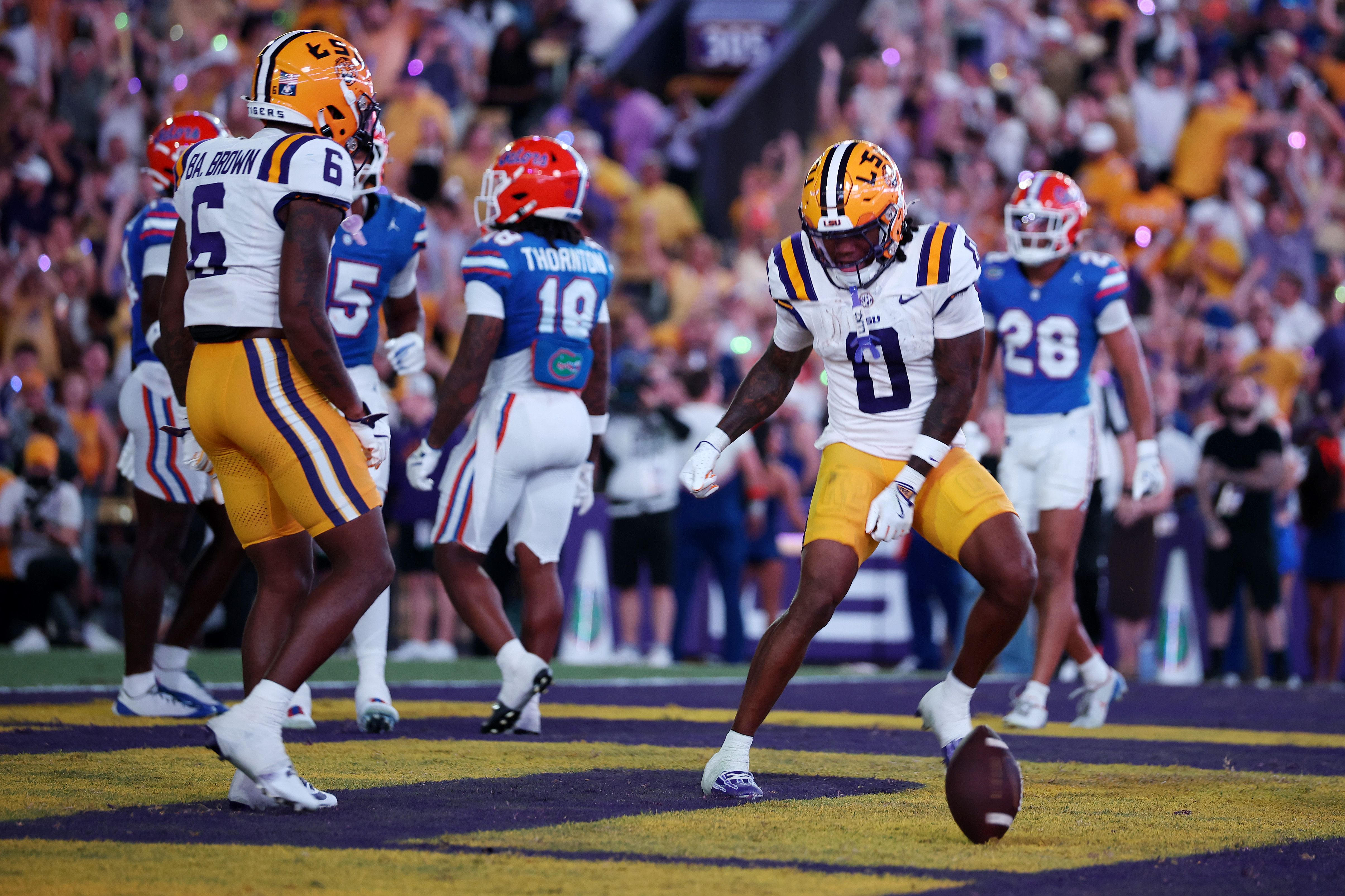 LSU Tigers players in white jerseys with purple and gold trim on a football field, facing Florida Gators in blue, with a rolling football on purple turf and a packed crowd in the background.