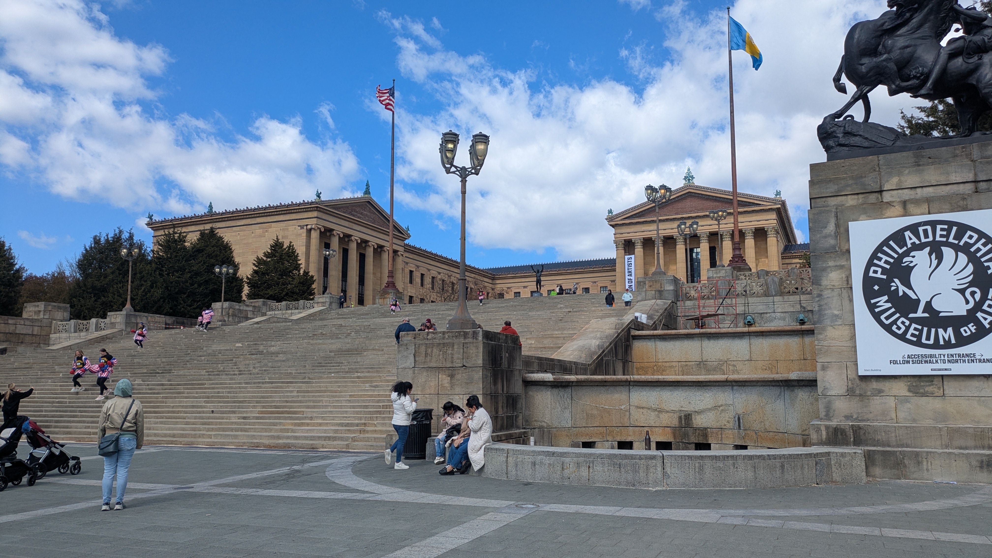 Blue sky over the Philadelphia Museum of Art steps, flanked by flagpoles and a large equestrian statue on the right. Visitors sit and walk on the wide stone staircase beneath the museum facade.