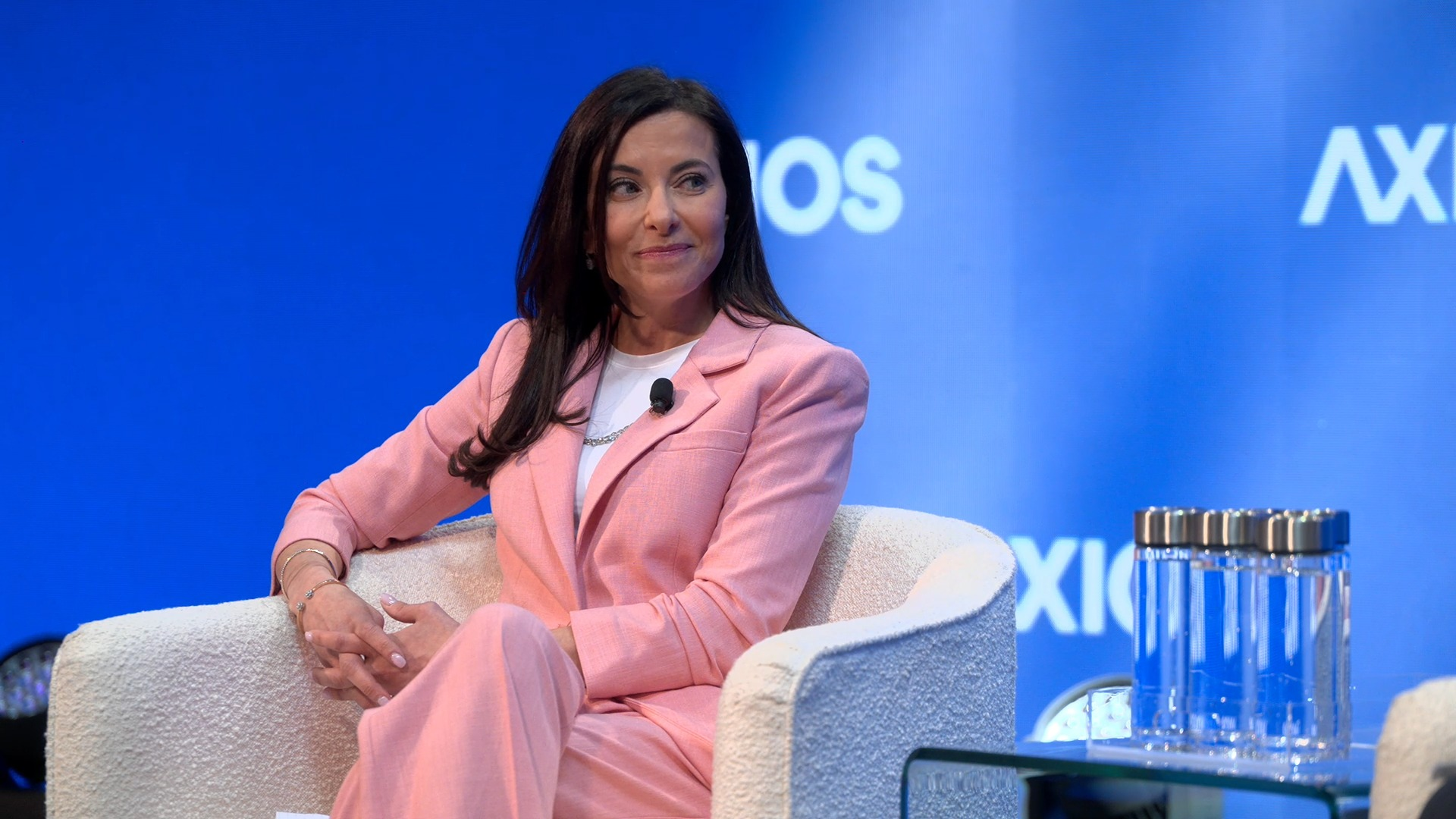 Woman in a pink suit sits on a cream textured armchair on a blue stage, wearing a white top and a lapel mic, with hands folded. A glass water jug sits on a glass table nearby.