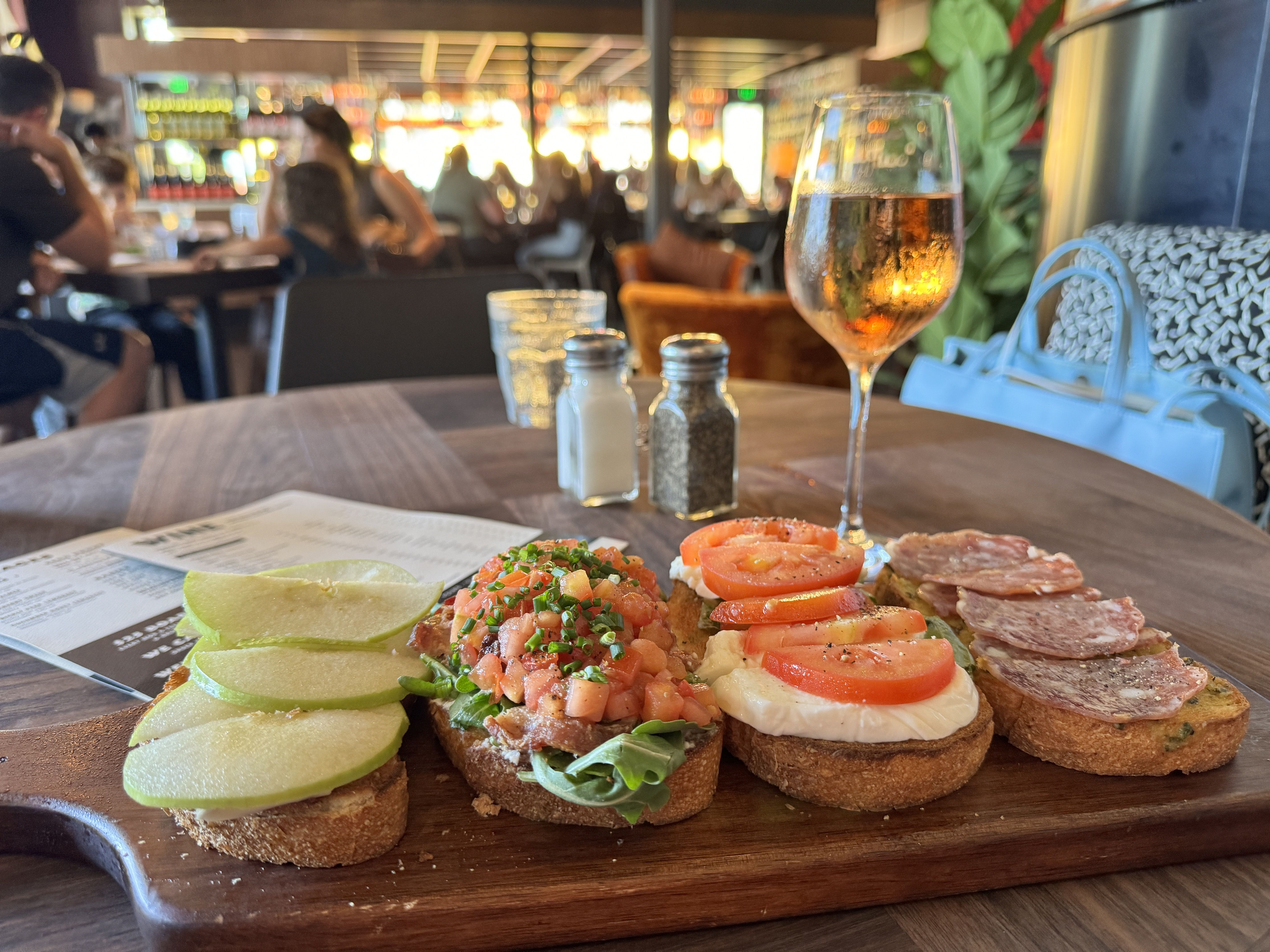 Crostini topped with chopped tomatoes and herbs on a wooden table, lime slices at side; a glass of wine and pepper shakers in foreground, diners blurred in a busy restaurant background.