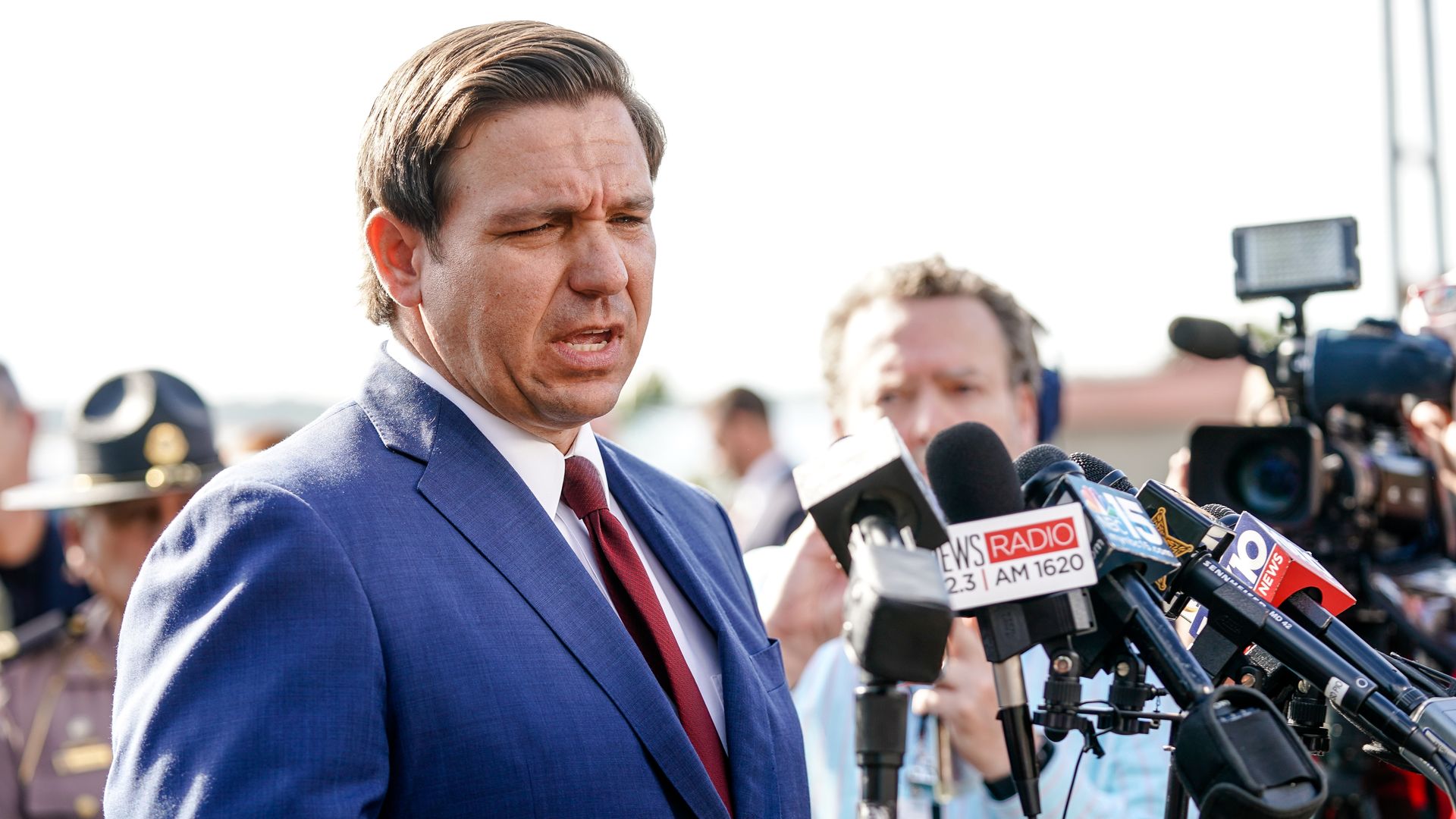 In this image, Ron DeSantis stands in a suit and tie and speaks into multiple reporters' microphones while standing outside.