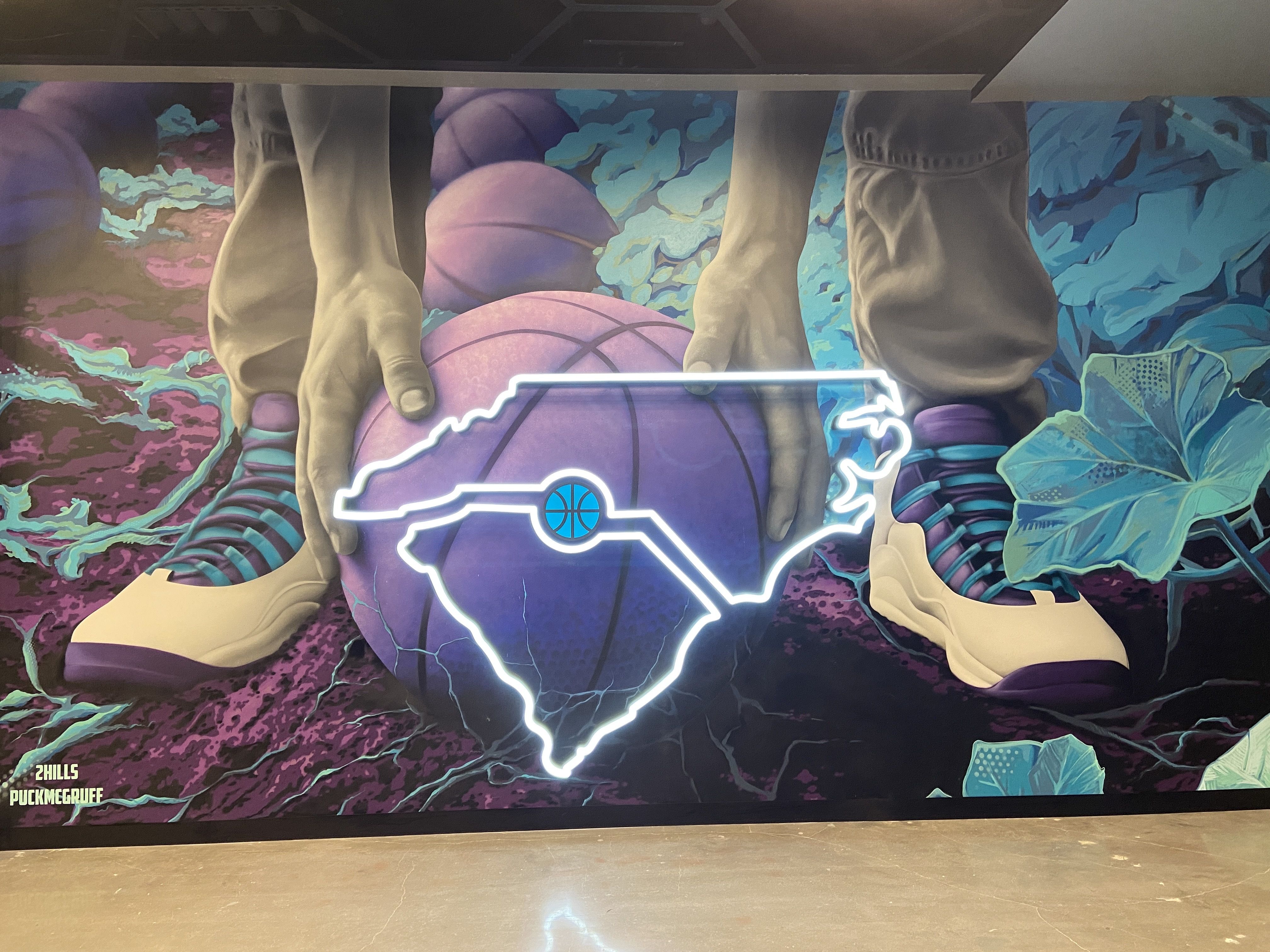 Mural showing hands holding a purple basketball behind an illuminated outline of the state of North Carolina with a blue basketball icon inside, set against purple and blue foliage.