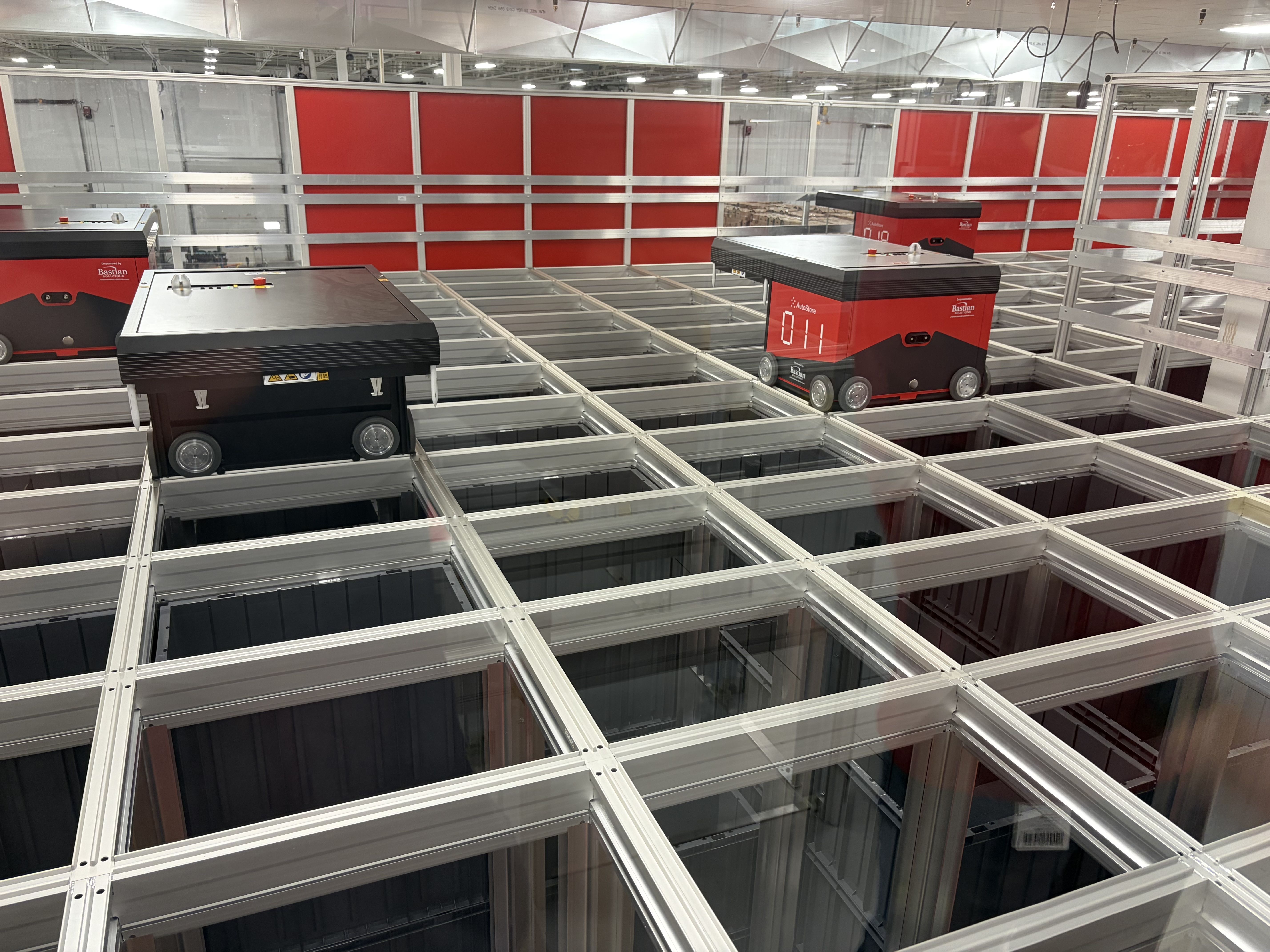 Industrial warehouse with red and black AutoStore storage robots on a white metal grid framework; red wall panels line the background under bright overhead lights.