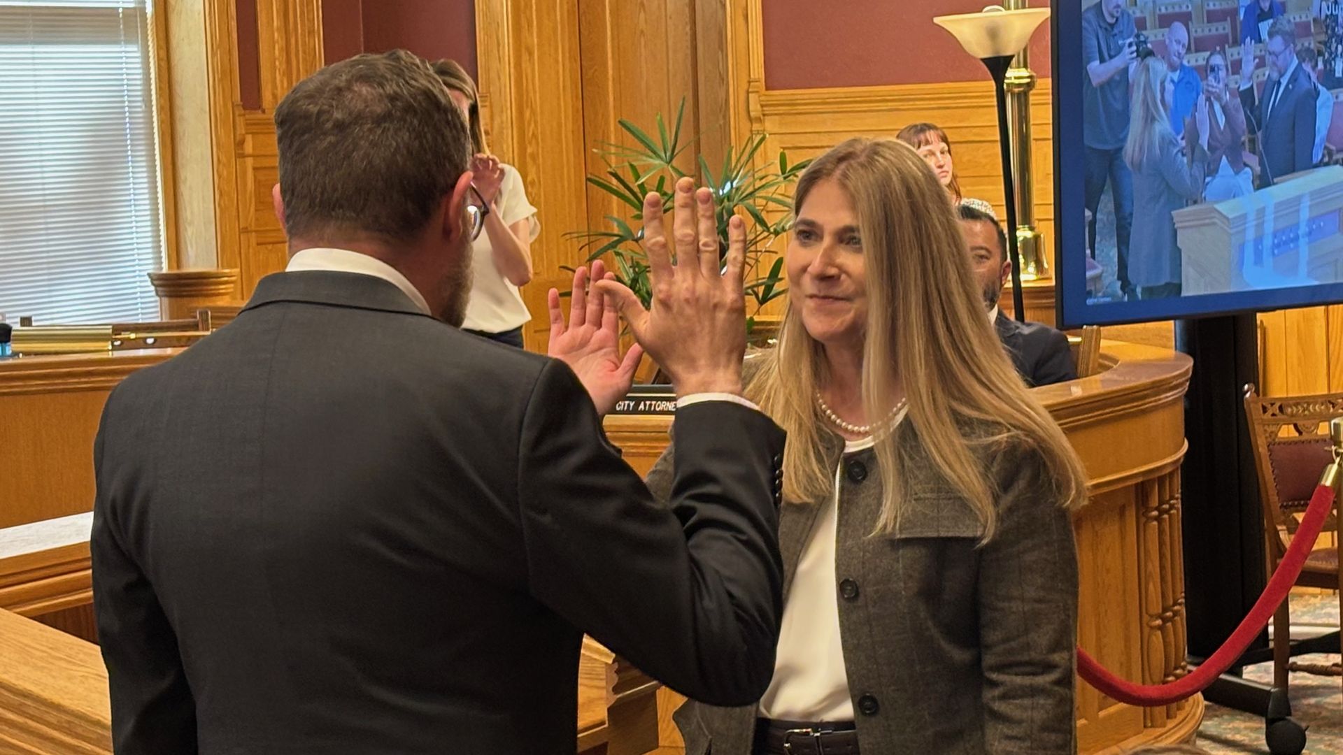 A man in a dark suit high-fives a blonde woman in a gray plaid blazer in a wood-paneled room; a large screen on the right shows people taking photos.