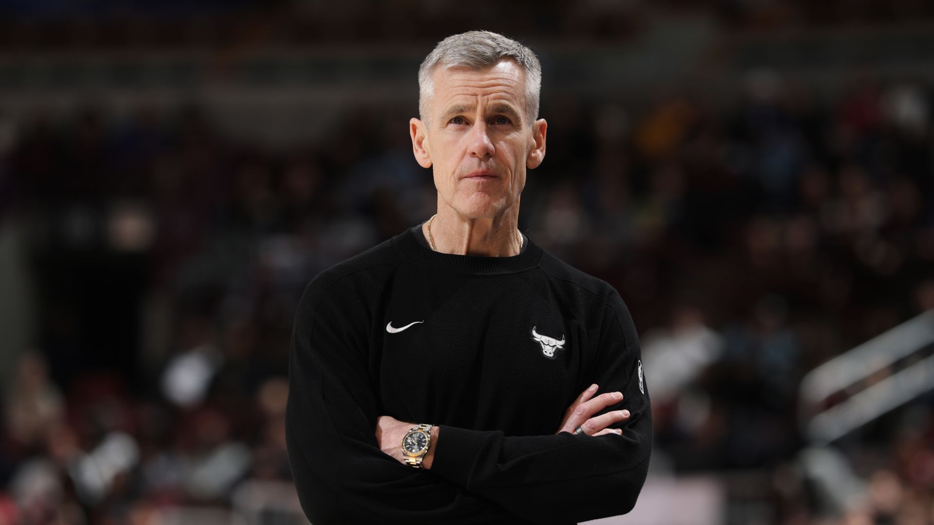 A man with short gray hair stands with arms crossed in a black Bulls sweatshirt with a Nike swoosh, in a crowded arena with a blurred audience.