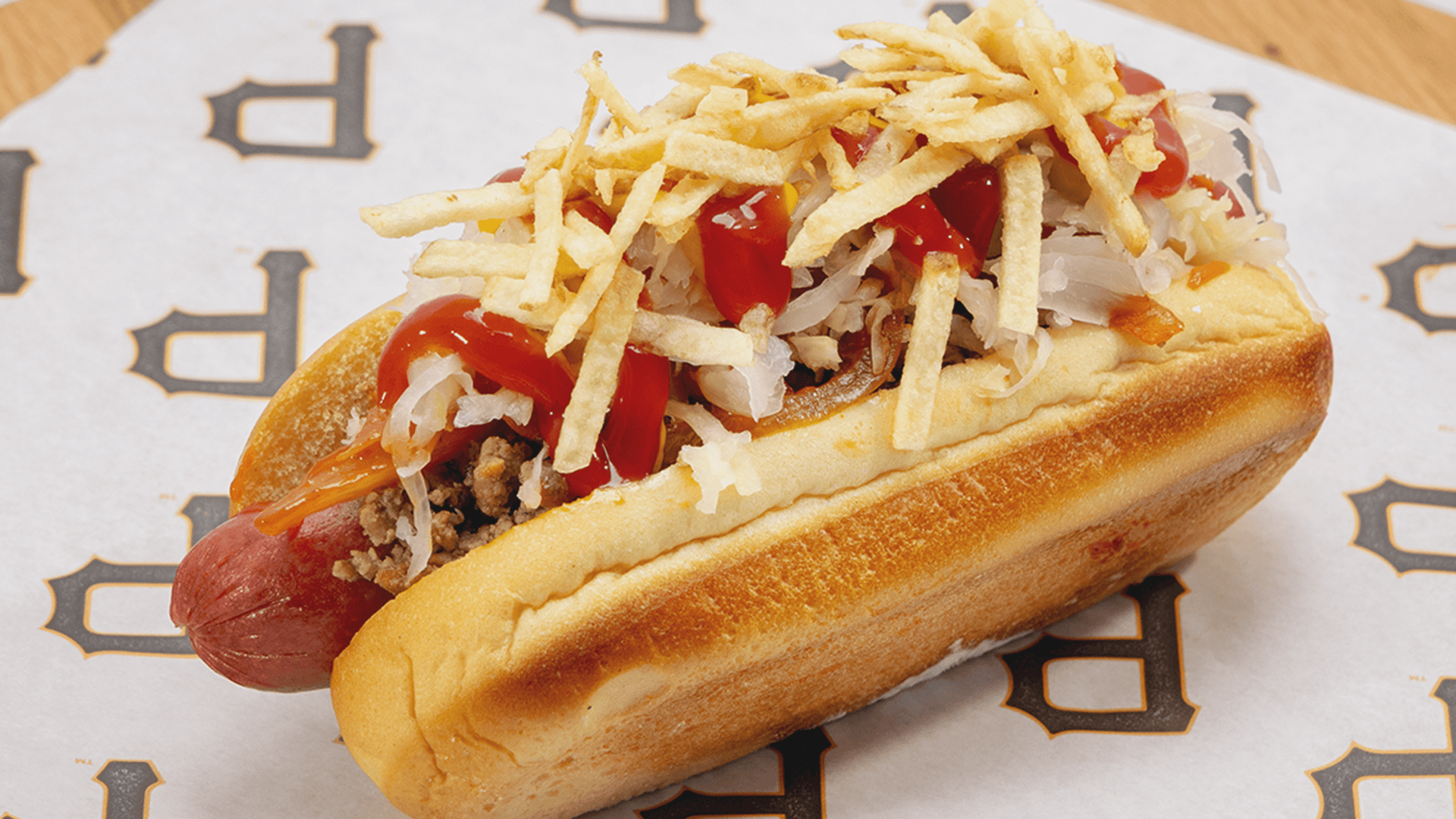 Toasted hot dog bun filled with sausage, ground meat, onions, red peppers, shredded cabbage, ketchup, and a mound of crispy shoestring potato sticks.