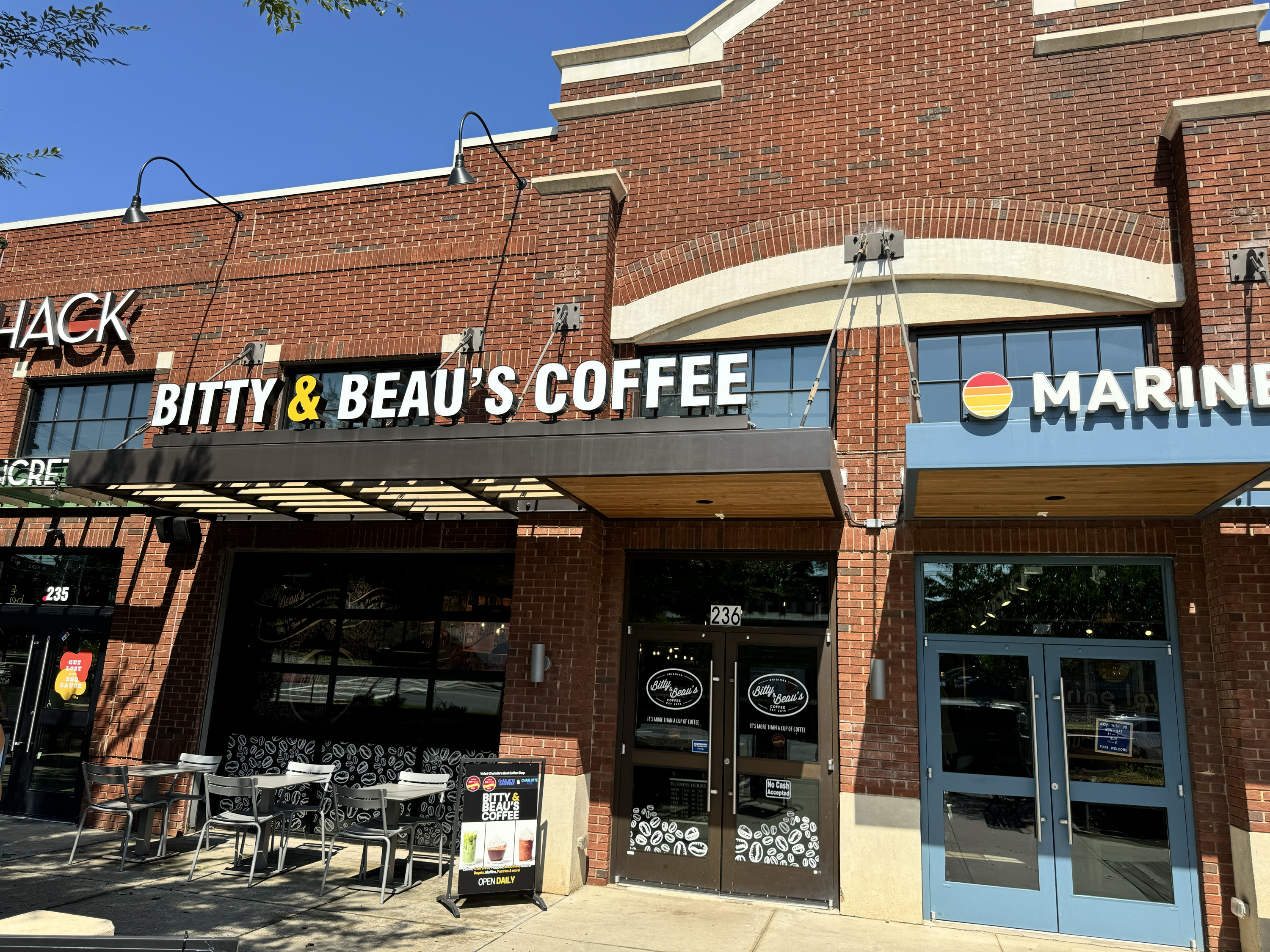 Brick storefronts with a cafe sign reading "BITTY & BEAU'S COFFEE", white letters with a yellow ampersand; blue sky, outdoor tables and chairs, and a neighboring shop with a round orange-yellow logo.