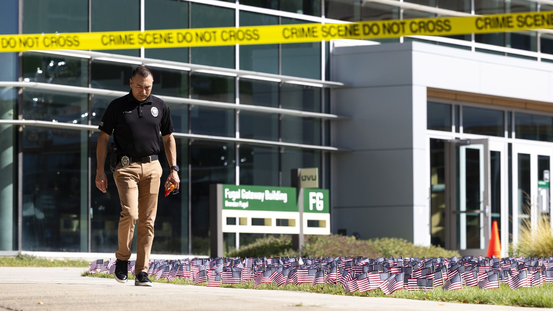 Police officer walking past yellow crime scene tape in front of a modern glass building with many small American flags planted in the grass near sign reading "Fugal Gateway Building" and "FG".