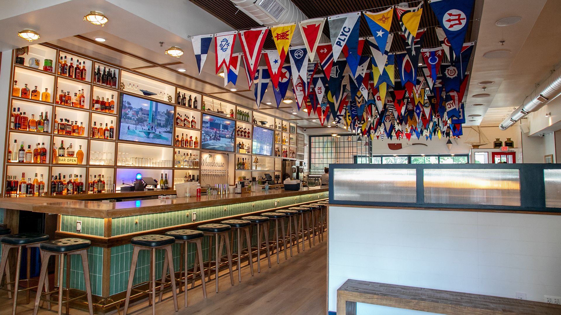 Bright, modern bar interior with a long wooden counter, green-tiled base, and tall stools. Bottles line illuminated shelves; nautical flags in red, blue, and white hang from the ceiling.