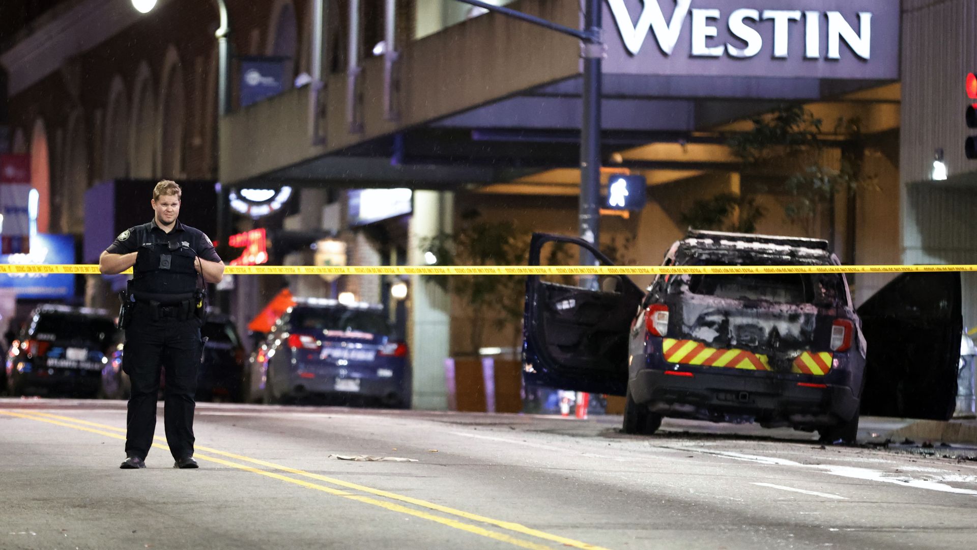 A police officer stands behind police tape and next to a burned-out police cruiser