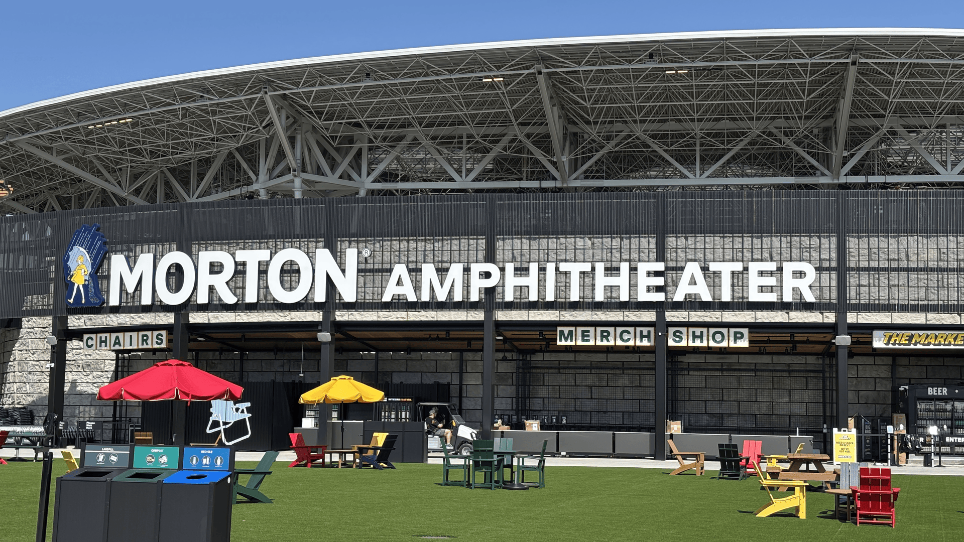 Exterior view of Morton Amphitheater featuring a large white sign reading "MORTON AMPHITHEATER" on a dark metal fence, with a latticed roof, green lawn, and colorful outdoor chairs and umbrellas in front.