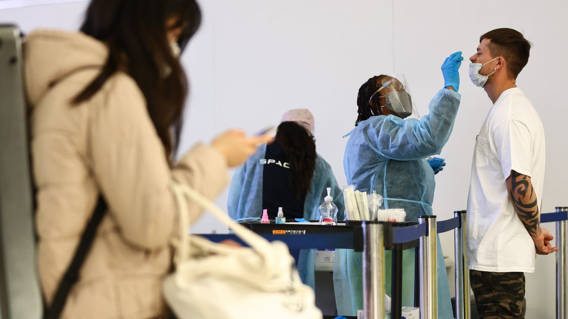 Someone dressed in PPE swabs another person's nose. A third person stands to the side of the image on their phone 