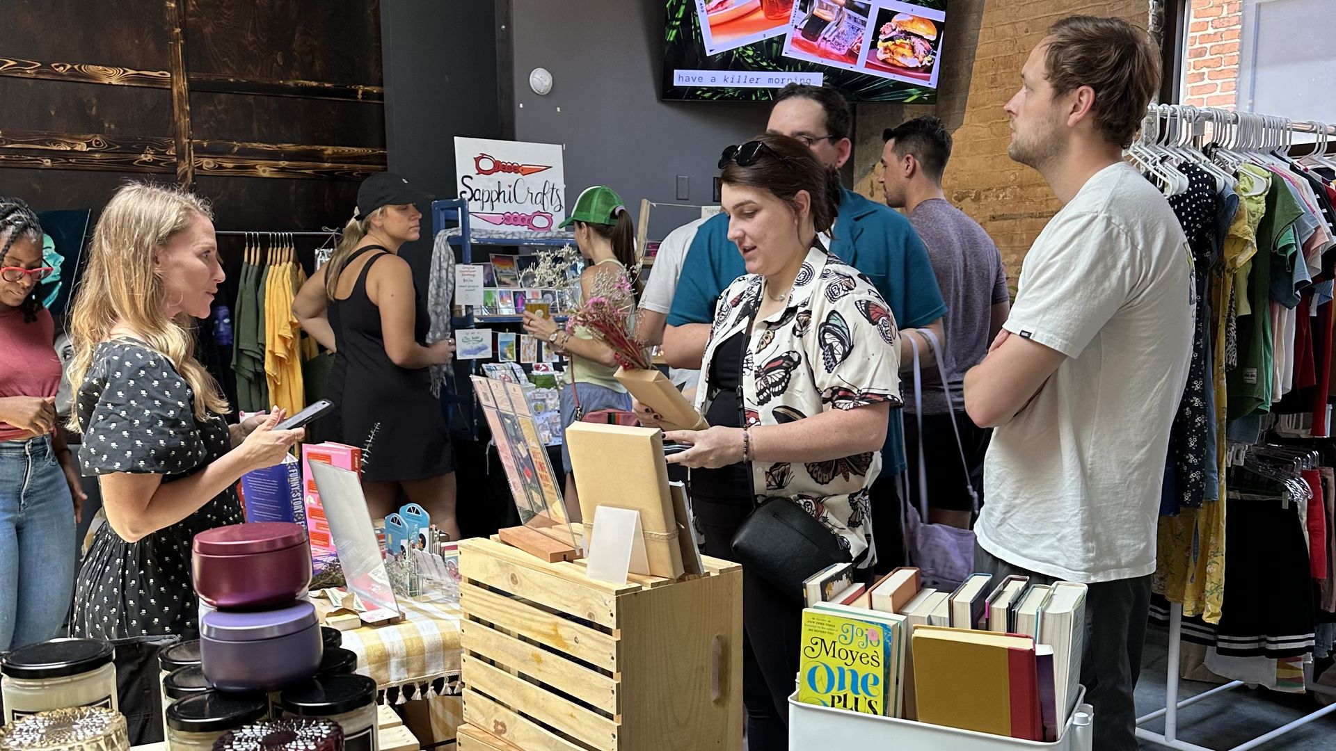 Indoor market scene with people browsing wooden displays of candles, books, and crafts, set against brick walls and a window letting in light, with a screen showing coffee images.